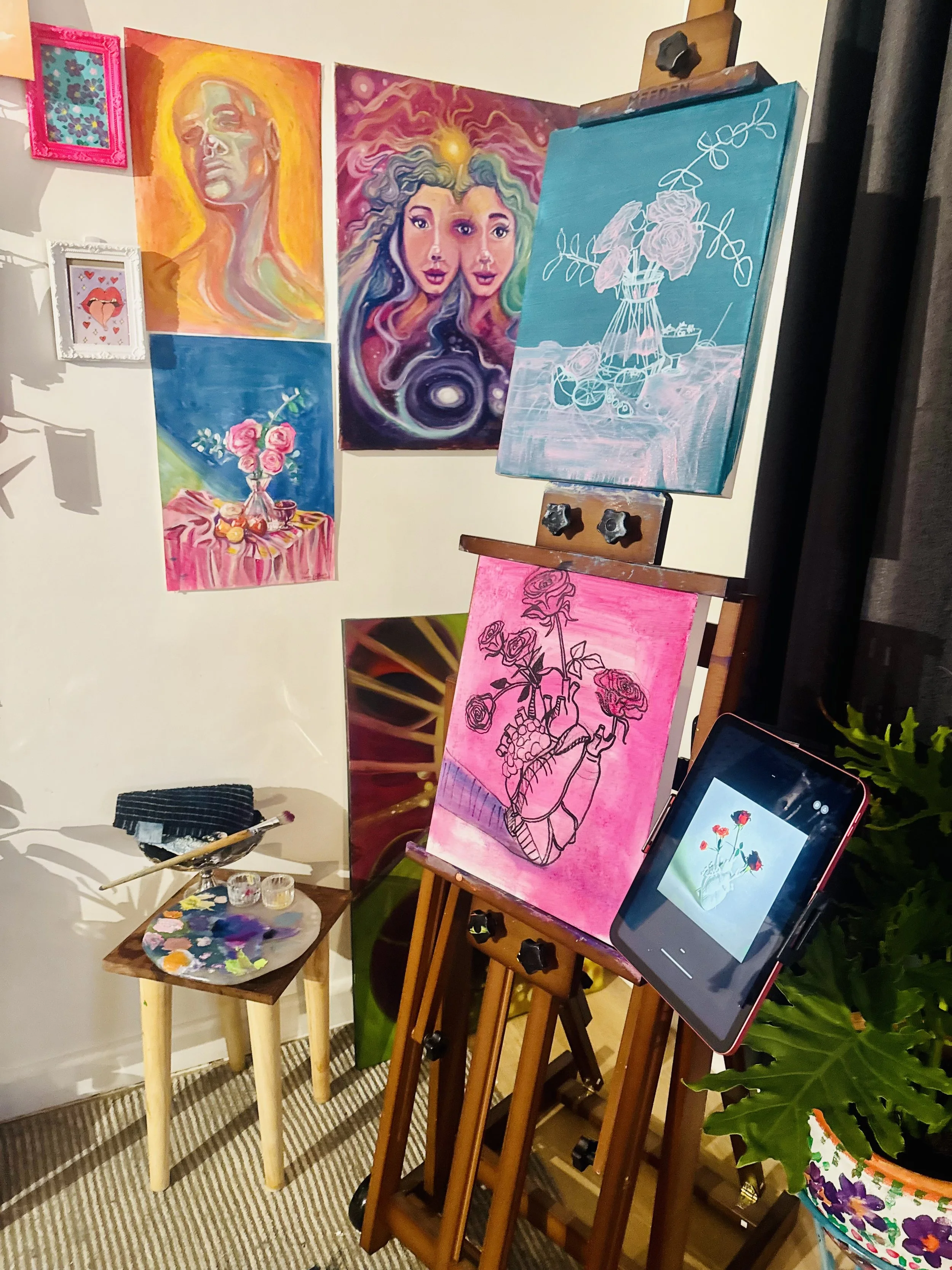 An art studio setup featuring multiple colorful paintings on the wall, an easel with a painting of a vase with roses, a tablet displaying an image of a vase with roses, and a small table with paint supplies. The paintings include abstract portraits, 