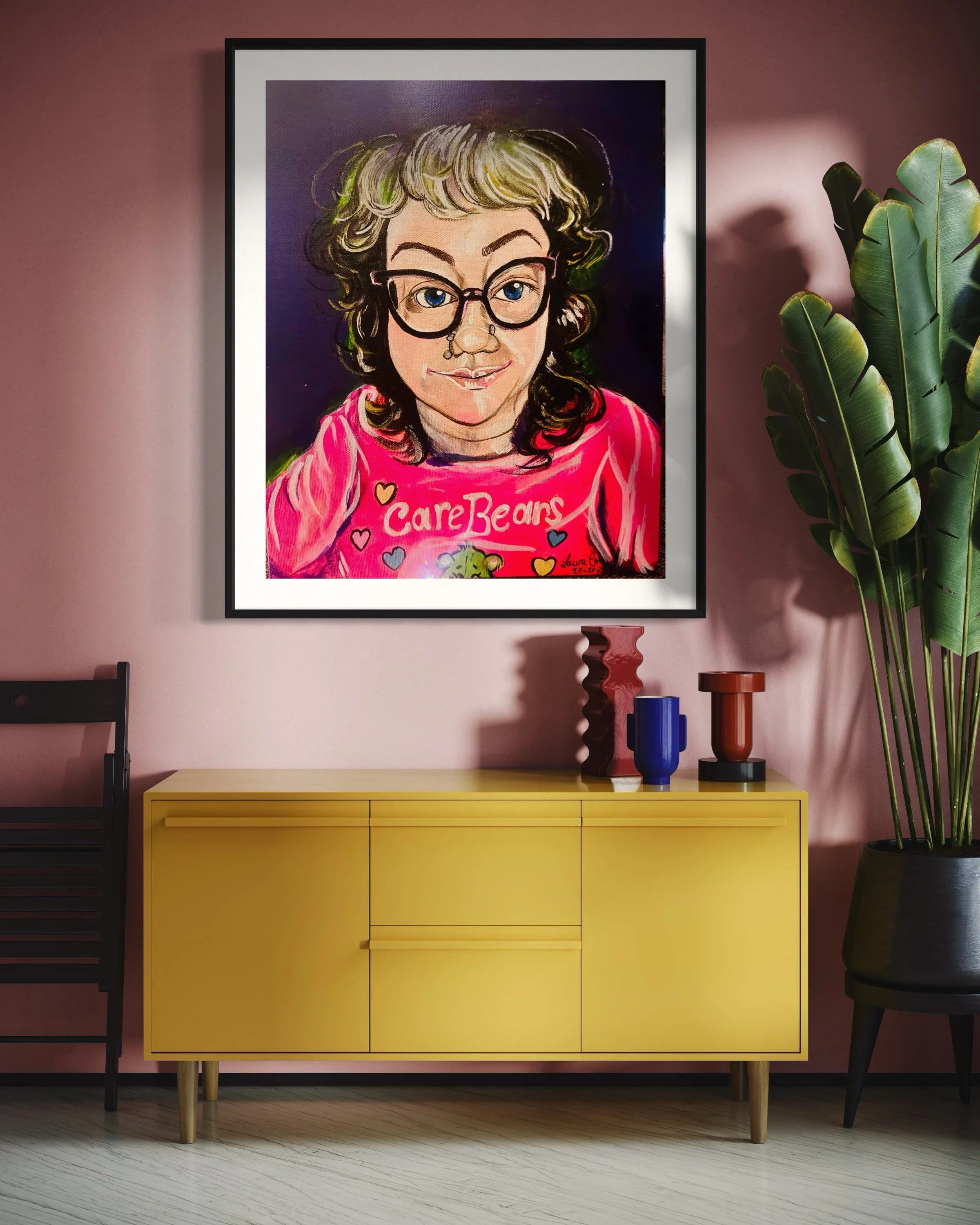 Colorful framed portrait of a woman with curly hair and glasses, wearing a pink Care Bears sweatshirt, hanging on a pink wall in a modern living room with a yellow sideboard, vases, and large green plant.