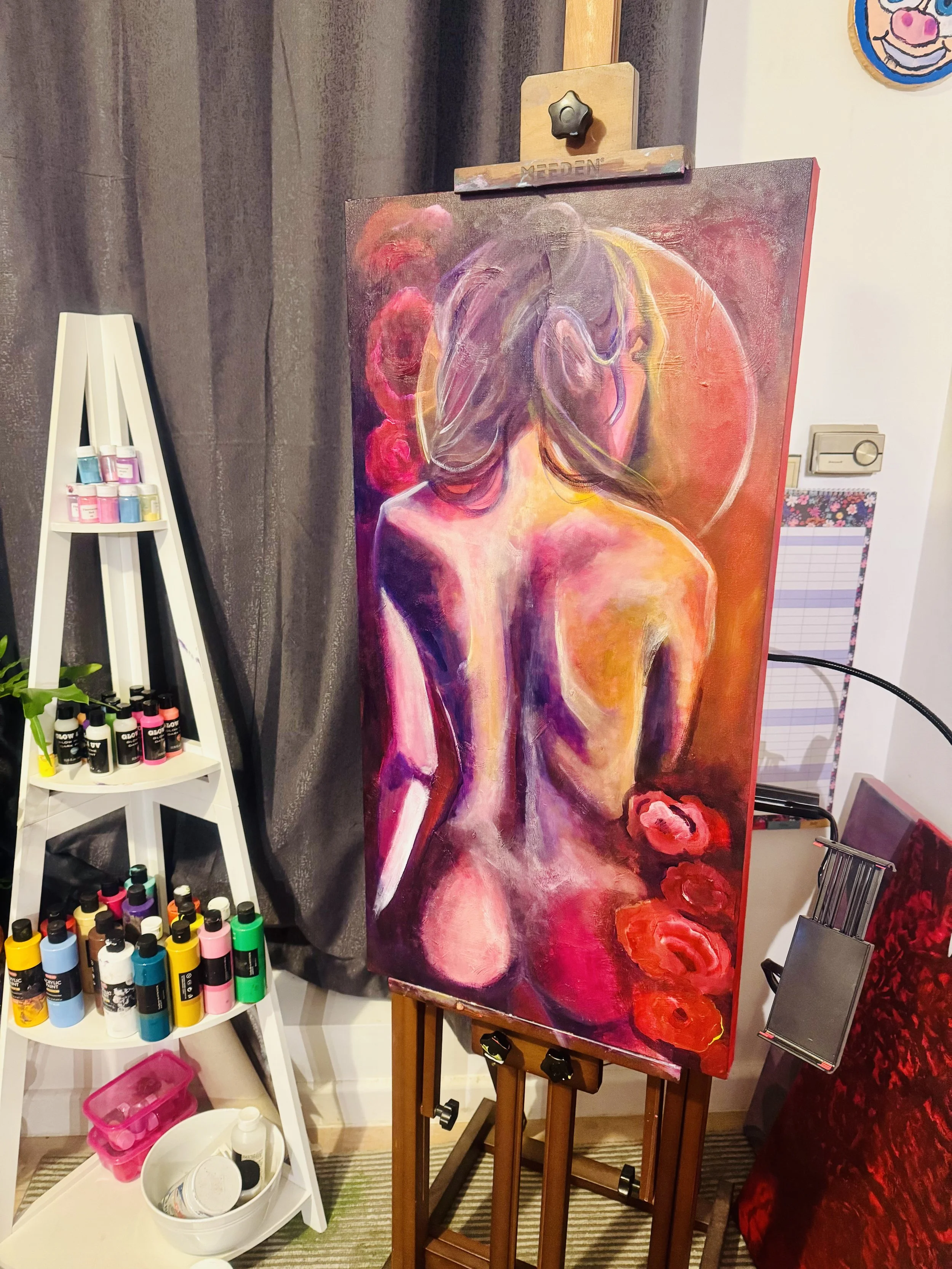 An abstract painting of a woman from behind, with her back and shoulders visible, surrounded by vibrant pink, purple, and red colors, on an easel in an art studio.