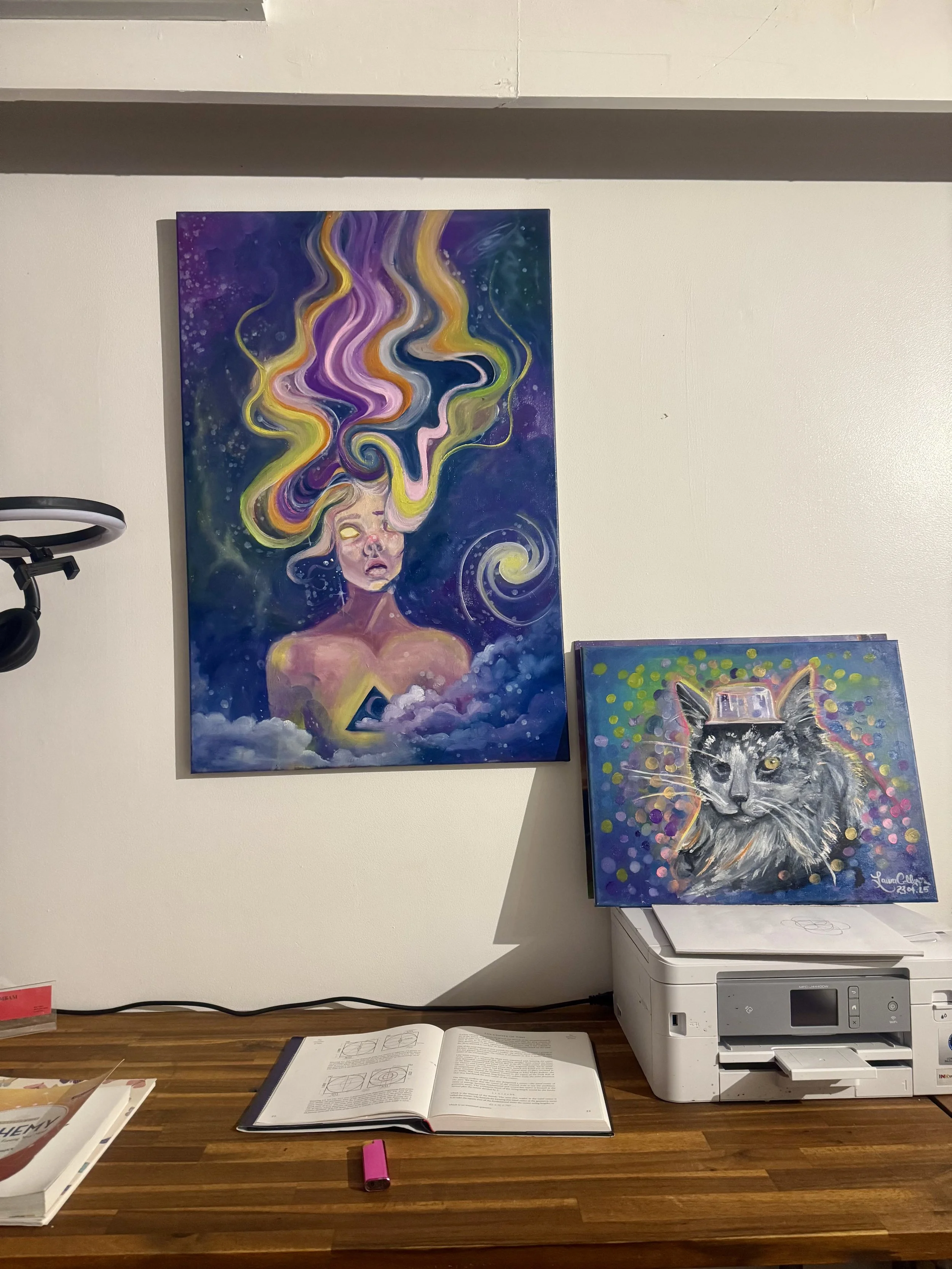 Two colorful paintings on a wall above a wooden desk. The larger painting features a surreal female figure with multicolored, flowing hair against a cosmic background. The smaller painting depicts a detailed portrait of a cat wearing a top hat, with 