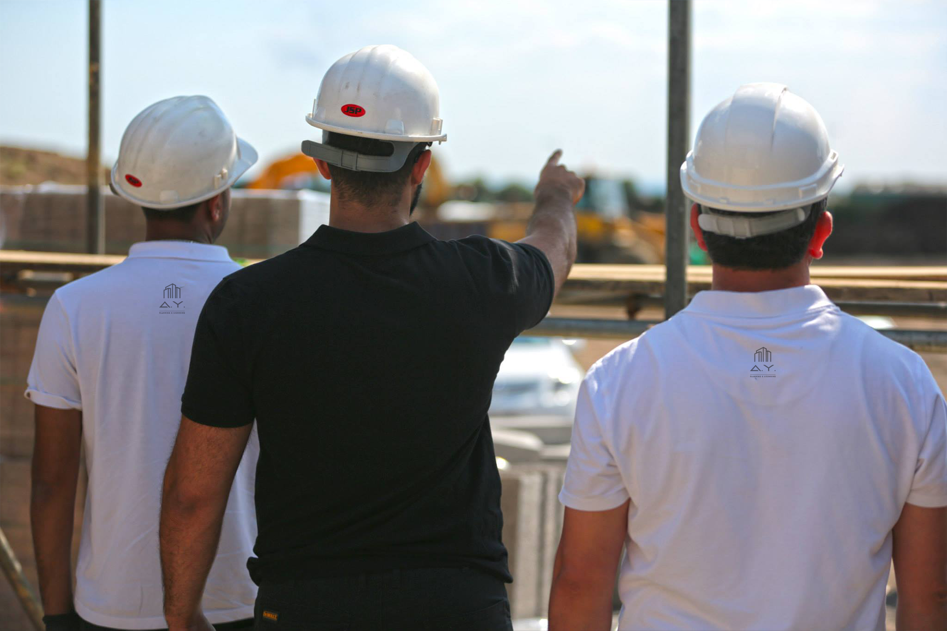 Three construction workers wearing white safety helmets, two men in white shirts and one man in a black shirt, are standing outdoors at a construction site, with the man in black pointing towards something in the distance.