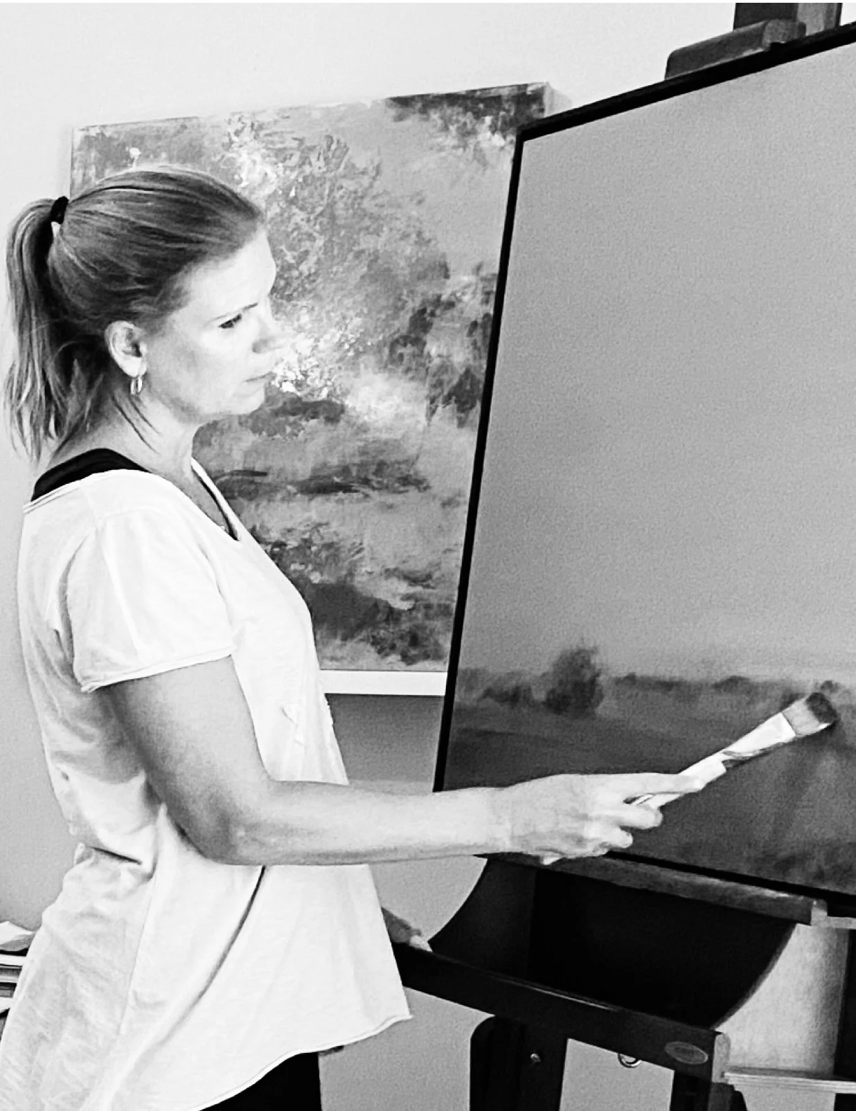 A woman with a ponytail standing next to an easel, holding a paintbrush, with an abstract landscape painting in the background.