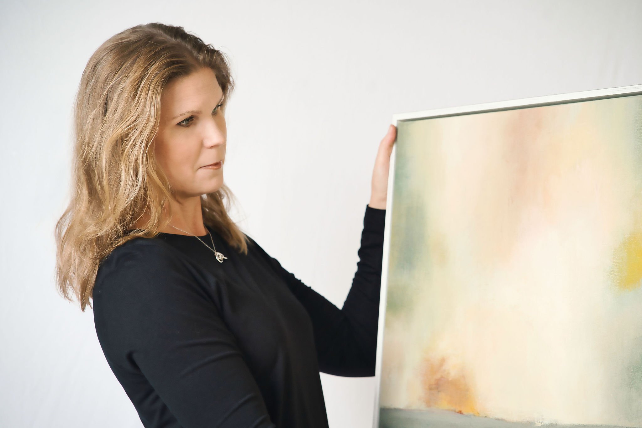 A woman with wavy blonde hair holding up a framed piece of abstract art.