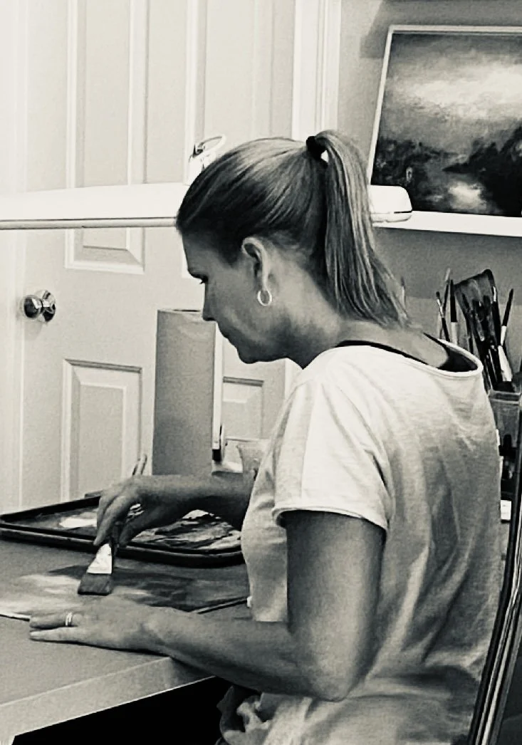 A woman sitting at a table, painting on a canvas with a paintbrush in her right hand, in a room with artwork and art supplies.