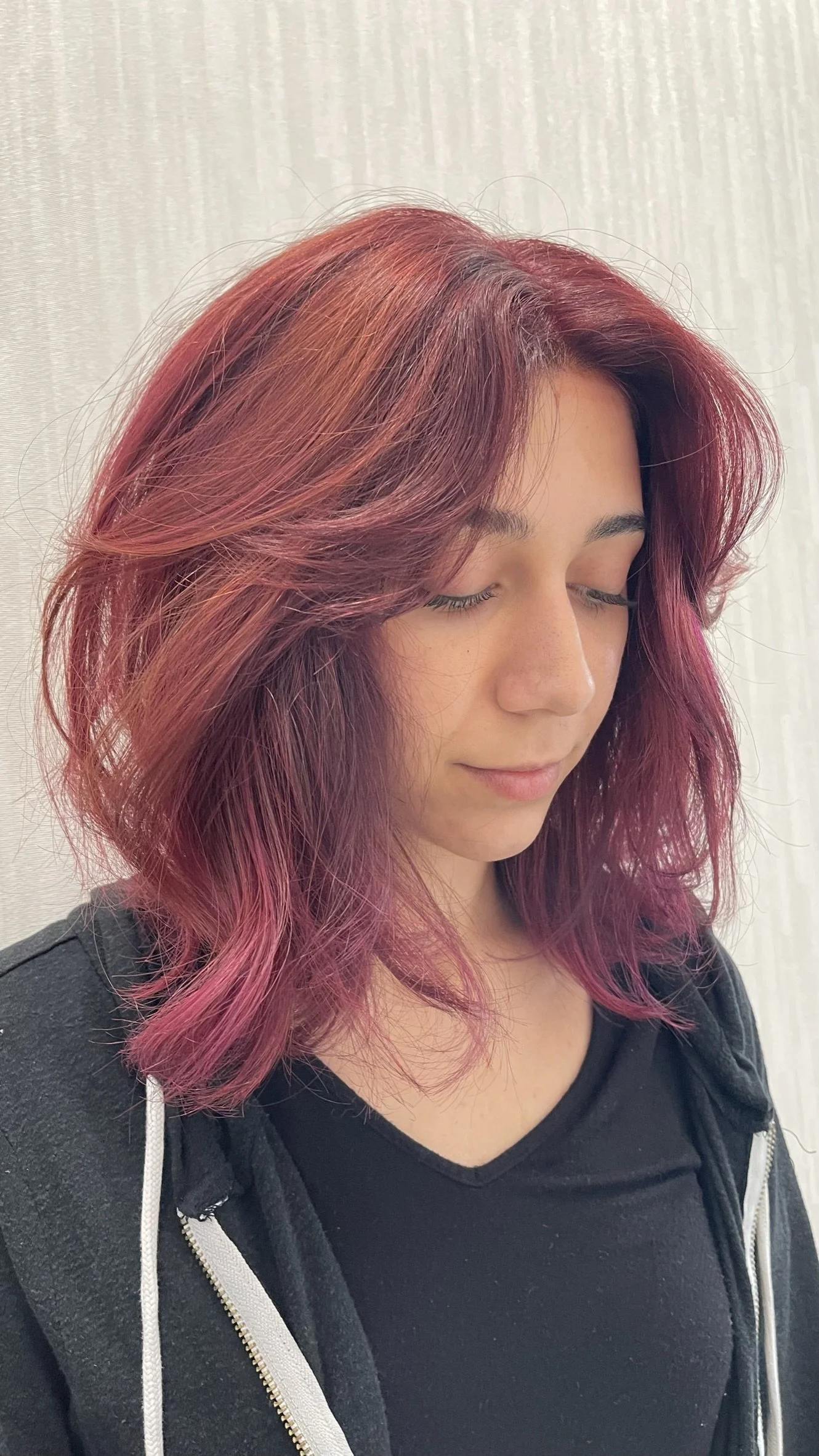 A woman with shoulder-length, wavy hair dyed in shades of pink and purple, with her eyes closed, standing in front of a light-colored curtain.