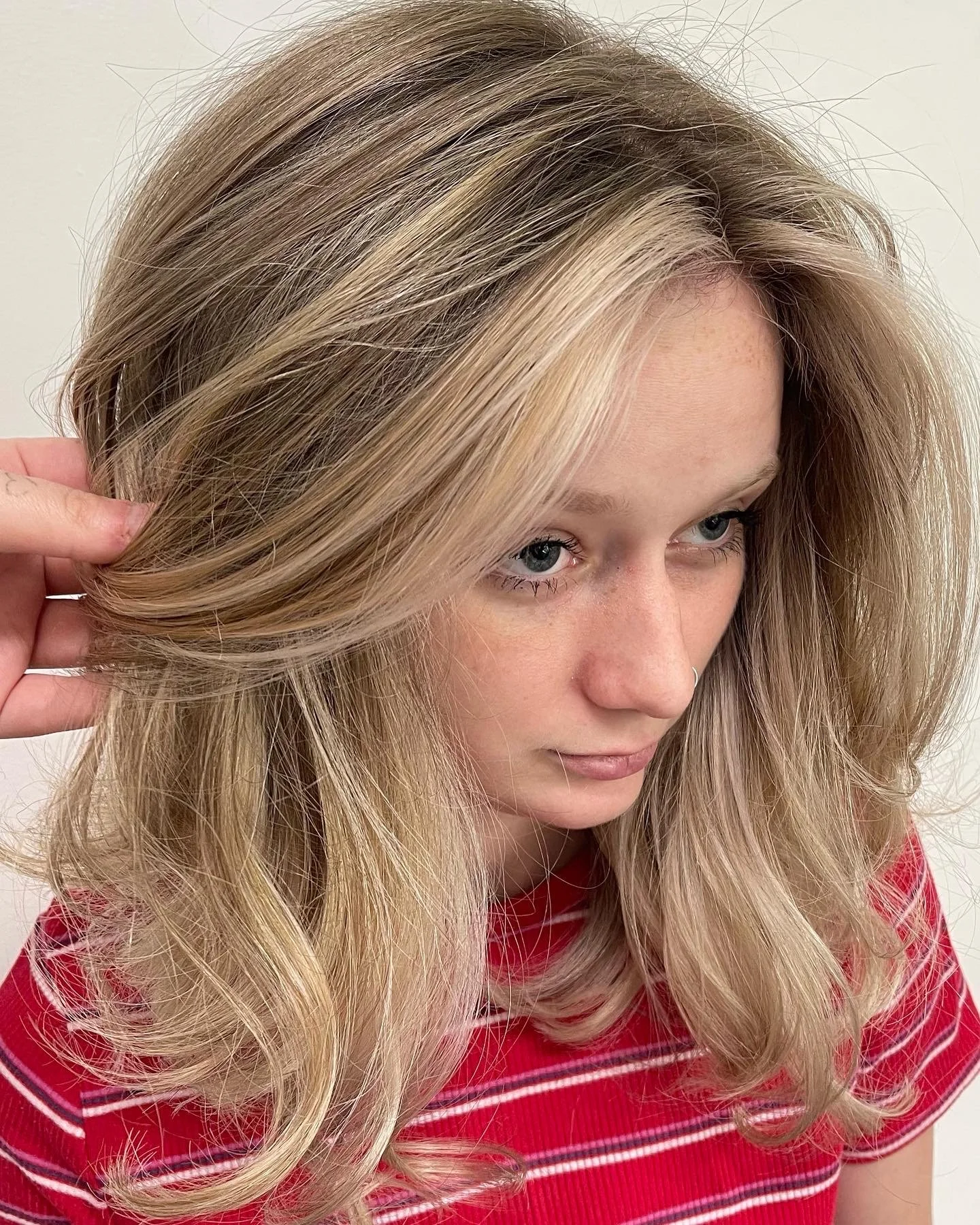 Close-up of a young woman with blonde, wavy hair and blue eyes, wearing a red striped shirt, touching her hair on the left side.