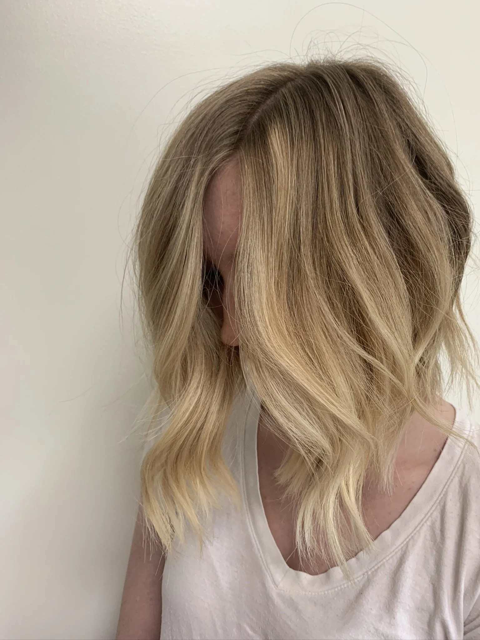 A woman with shoulder-length blonde hair styled in loose waves, wearing a white top, standing against a plain white wall.