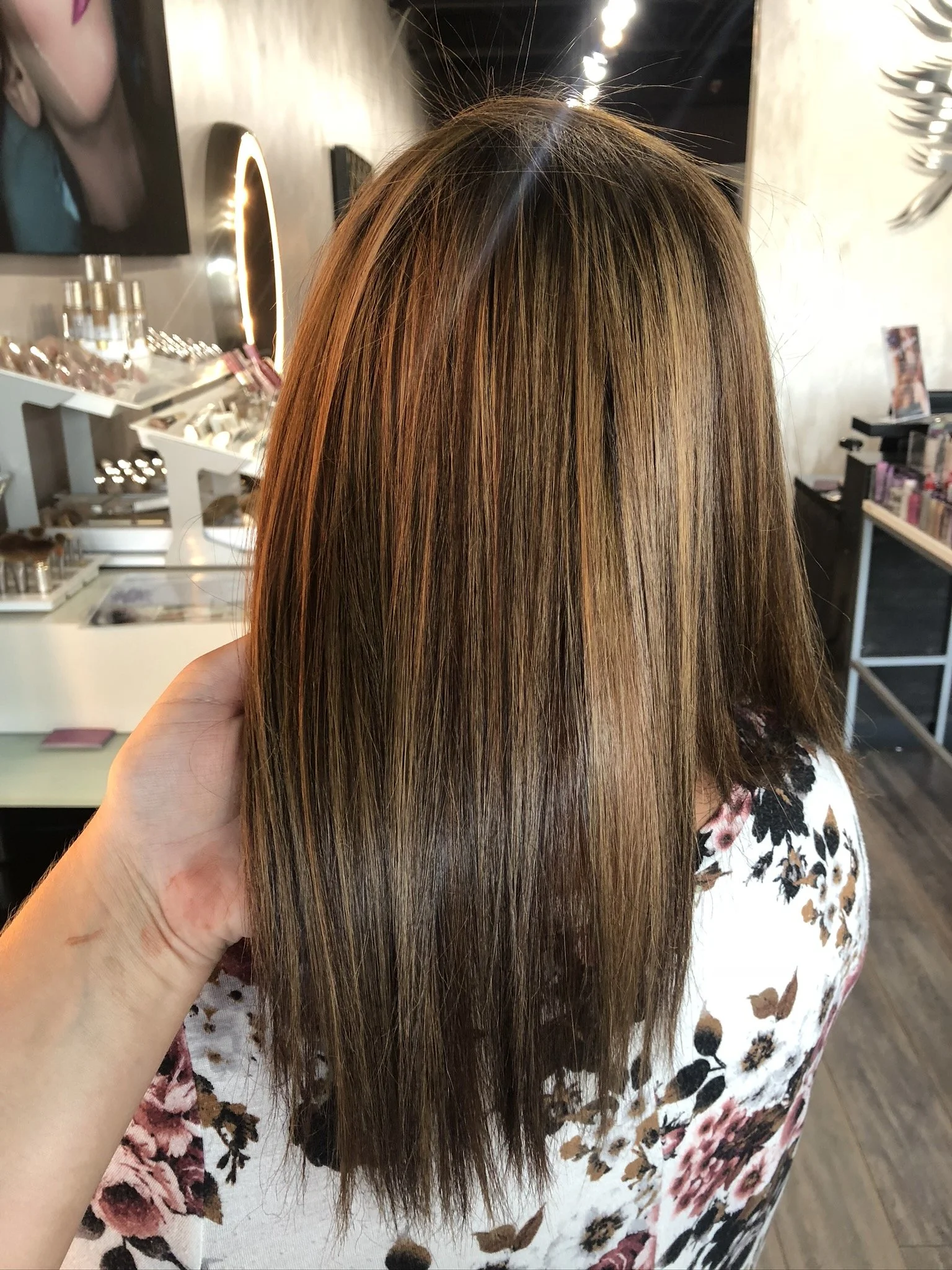 A woman with straight, shoulder-length hair that is a blend of brown and blonde highlights, sitting in a salon with makeup and hair products visible in the background.
