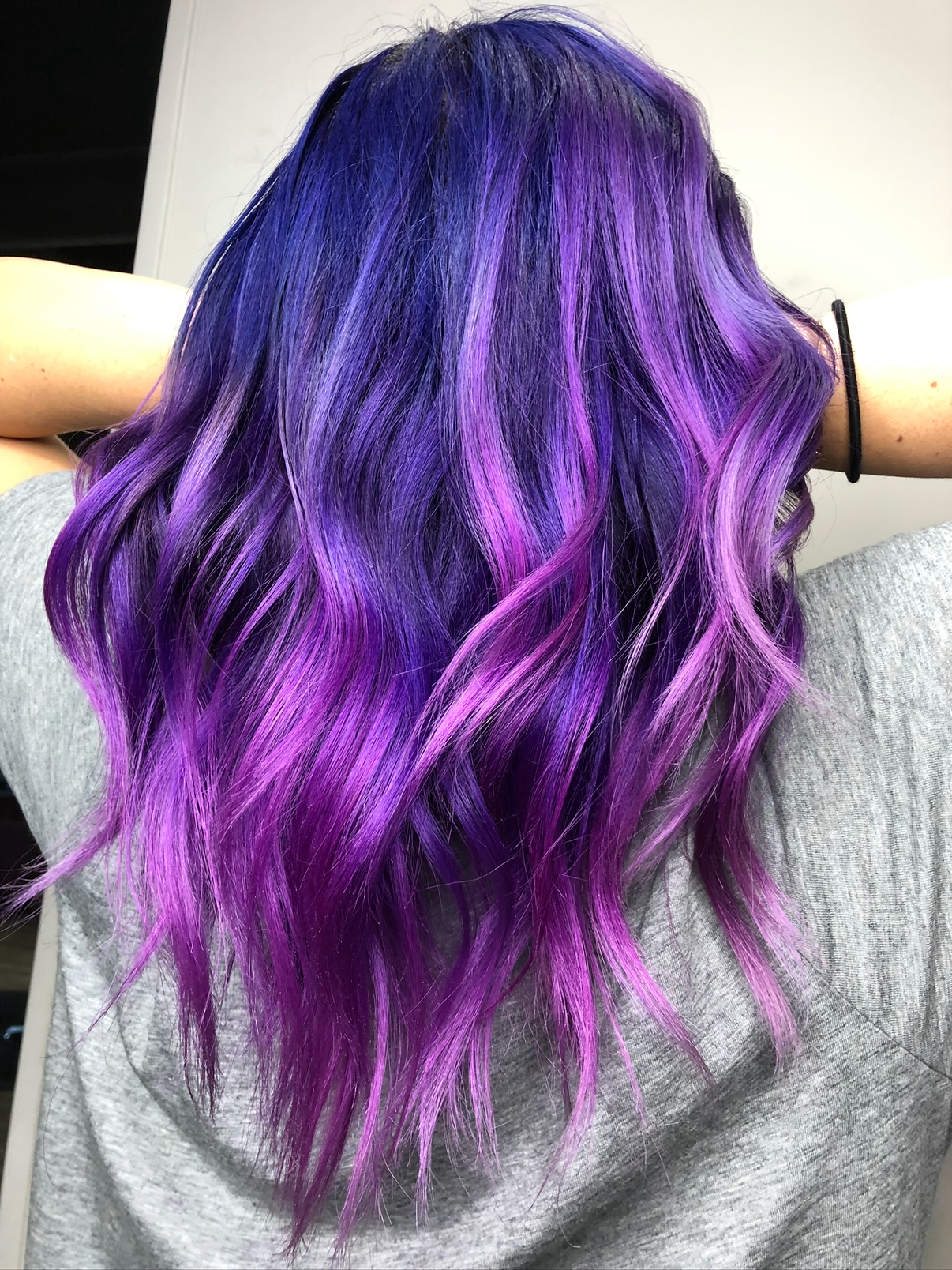 Back of a person's head with wavy, purple and pink ombre hair, wearing a gray hoodie and a black bracelet.