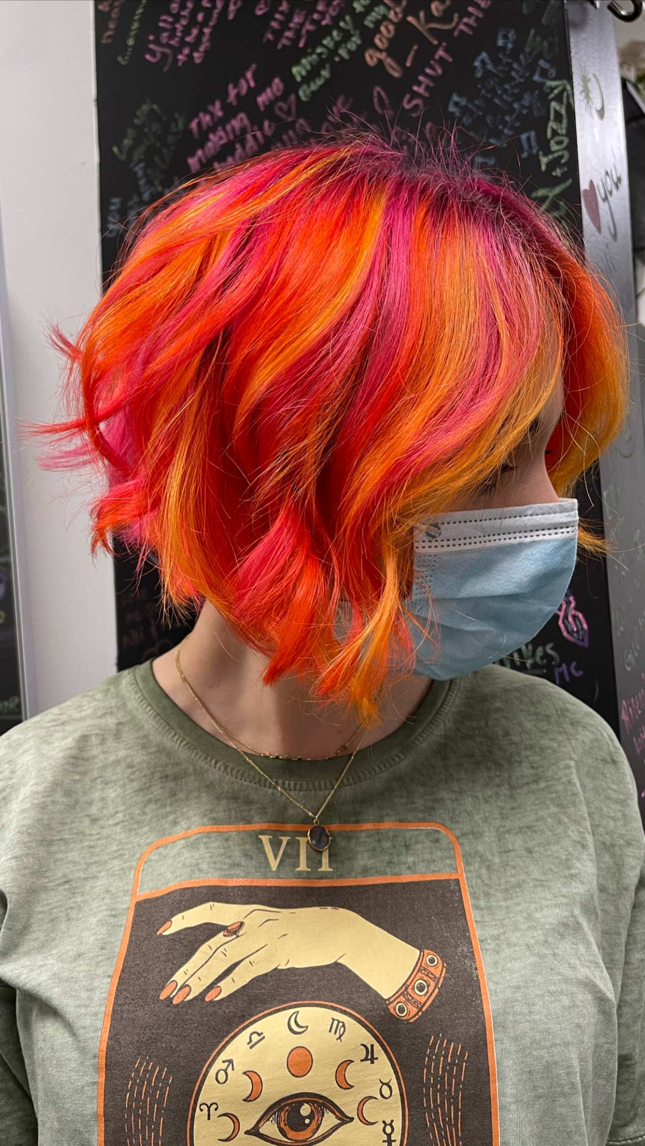 A person with a vibrant, multicolored, wavy bob haircut in shades of red, orange, pink, and yellow, wearing a light green sweatshirt with a graphic design, layered necklaces, and a blue face mask, standing in front of a black chalkboard with colorful