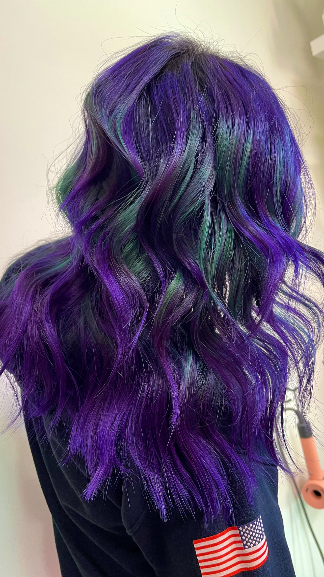 Back view of a person with wavy, shoulder-length hair dyed in purple, green, and blue colors, with a small American flag patch on their clothing.