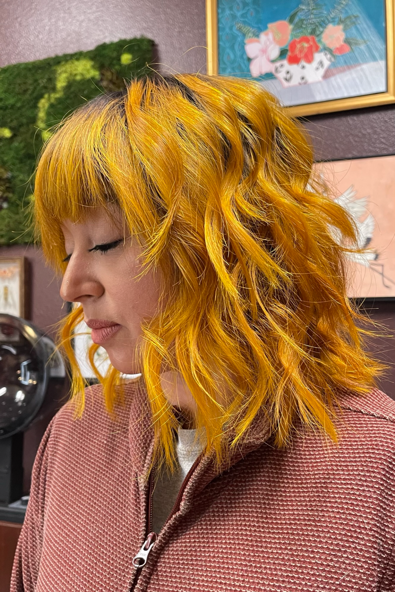 A woman with curly bright yellow and orange hair, seen in profile, wearing a pink textured zip-up jacket, in an indoor setting with various artwork and a green moss wall in the background.