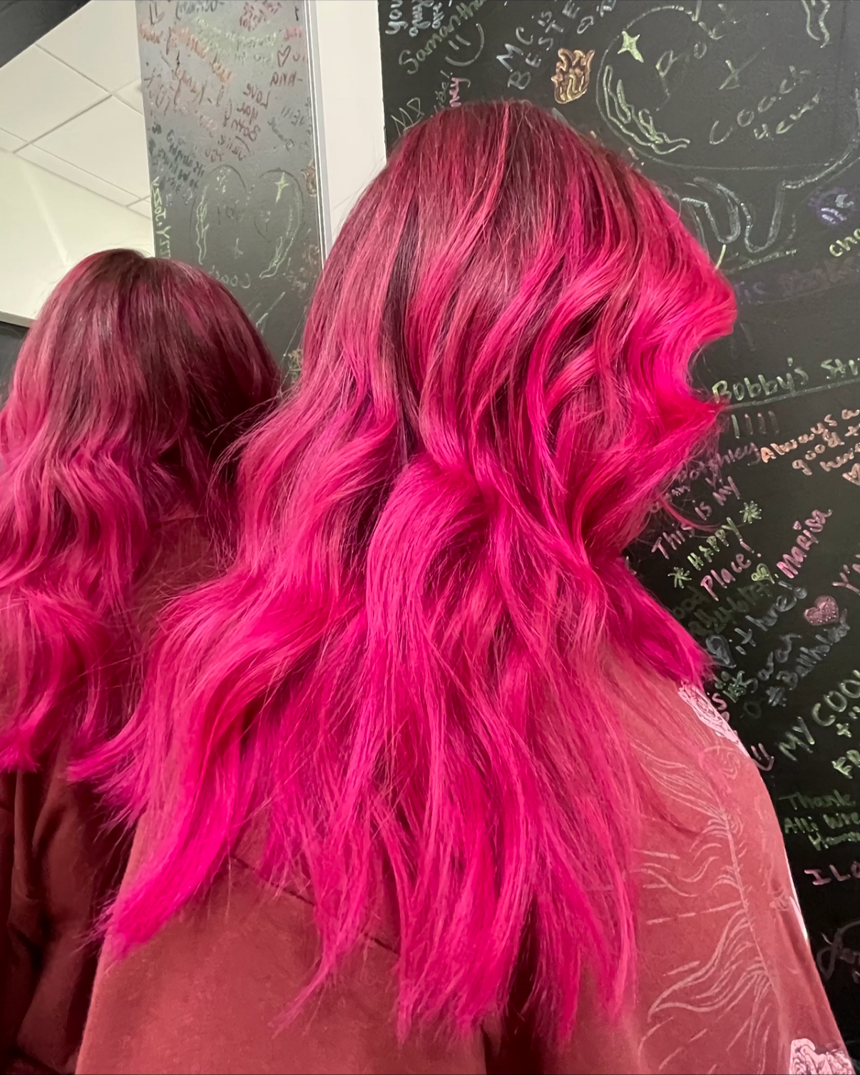 Woman with long wavy hair dyed bright pink, standing in front of a black chalkboard covered with colorful handwritten notes and doodles.