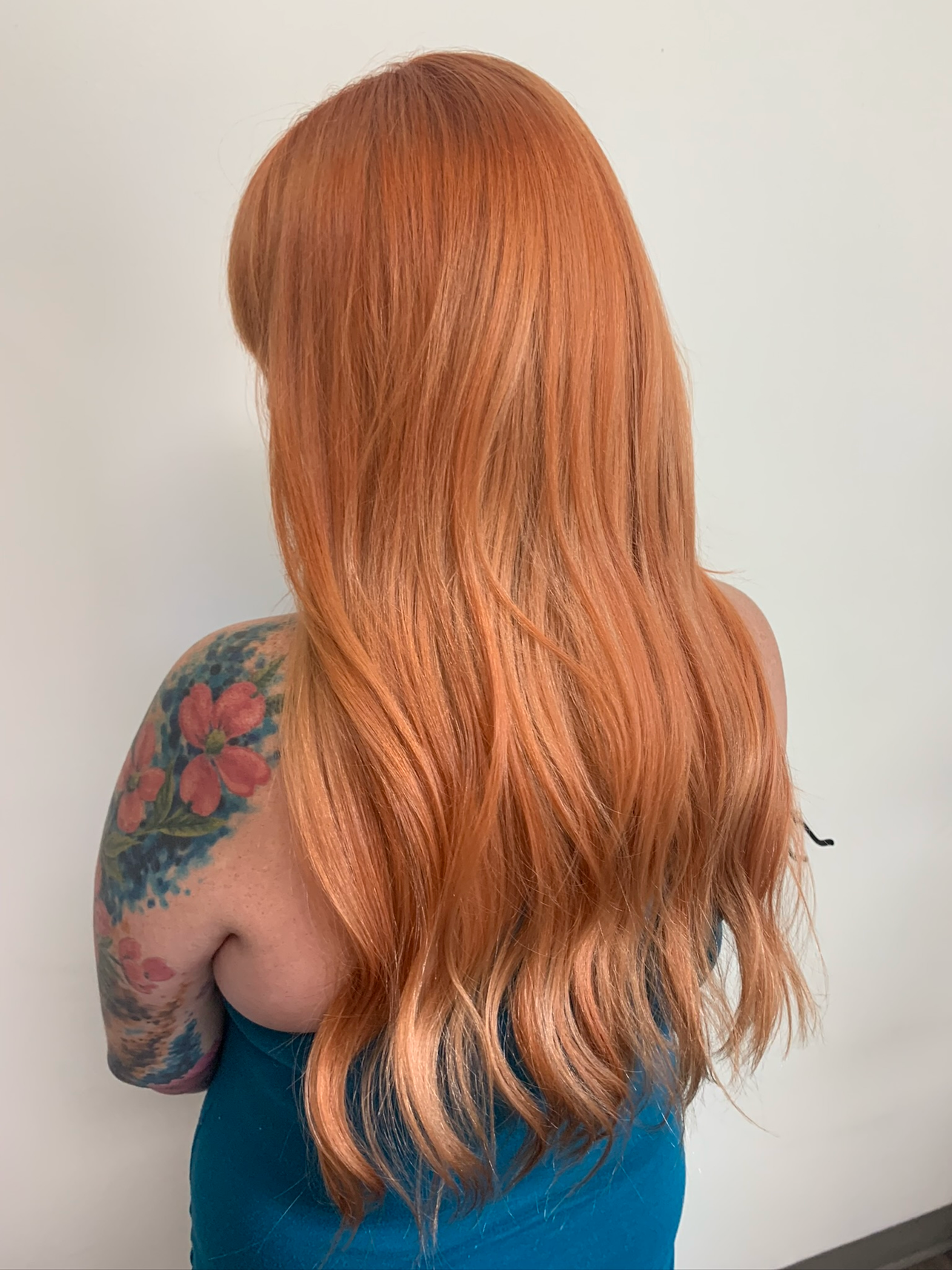 Back view of a woman with long, wavy, orange-red hair and a colorful floral tattoo on her left shoulder, wearing a teal top, against a plain white background.