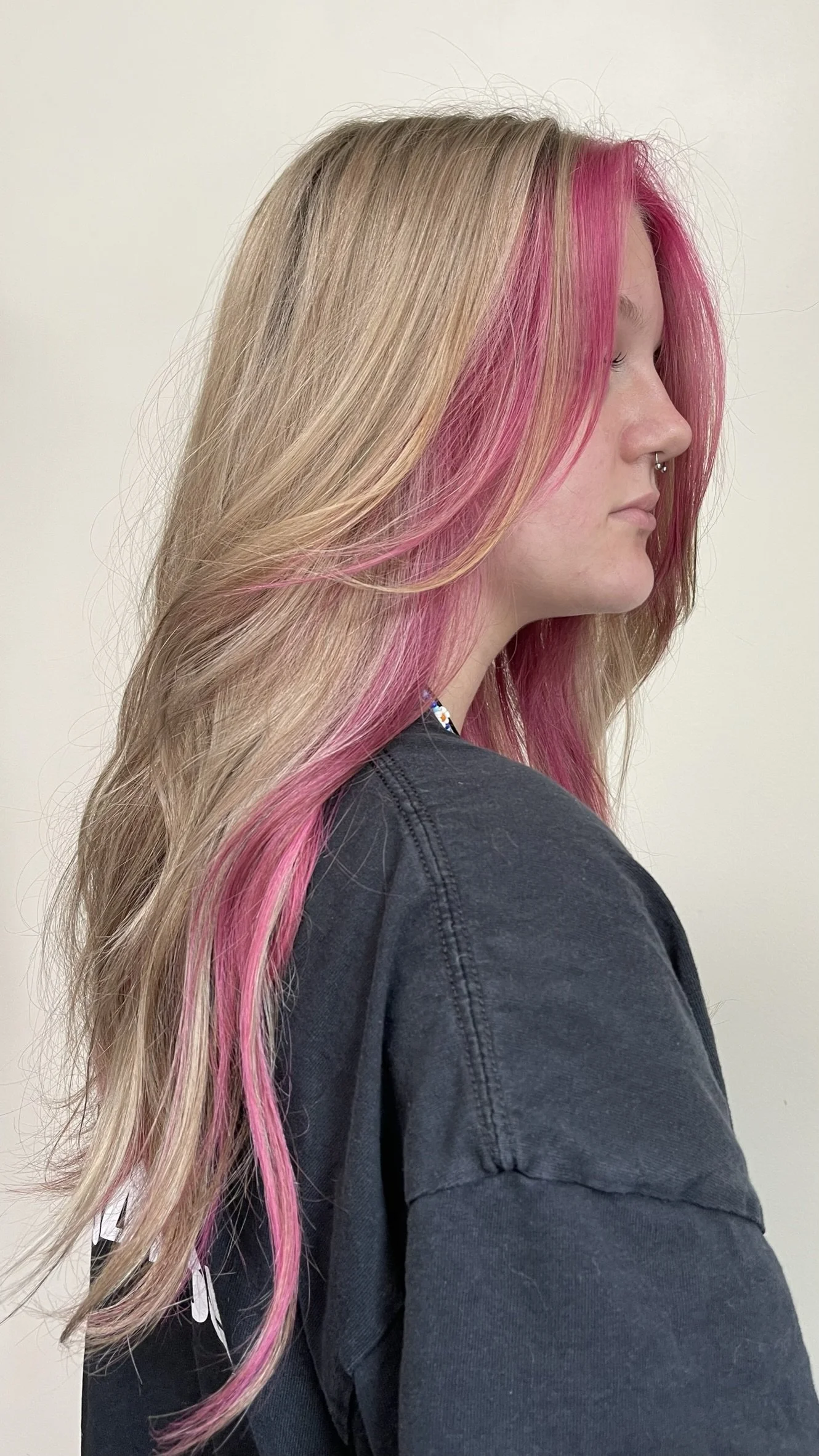 Side profile of a woman with long, blonde hair that has streaks of pink toward the front, wearing a dark gray shirt against a plain white background.