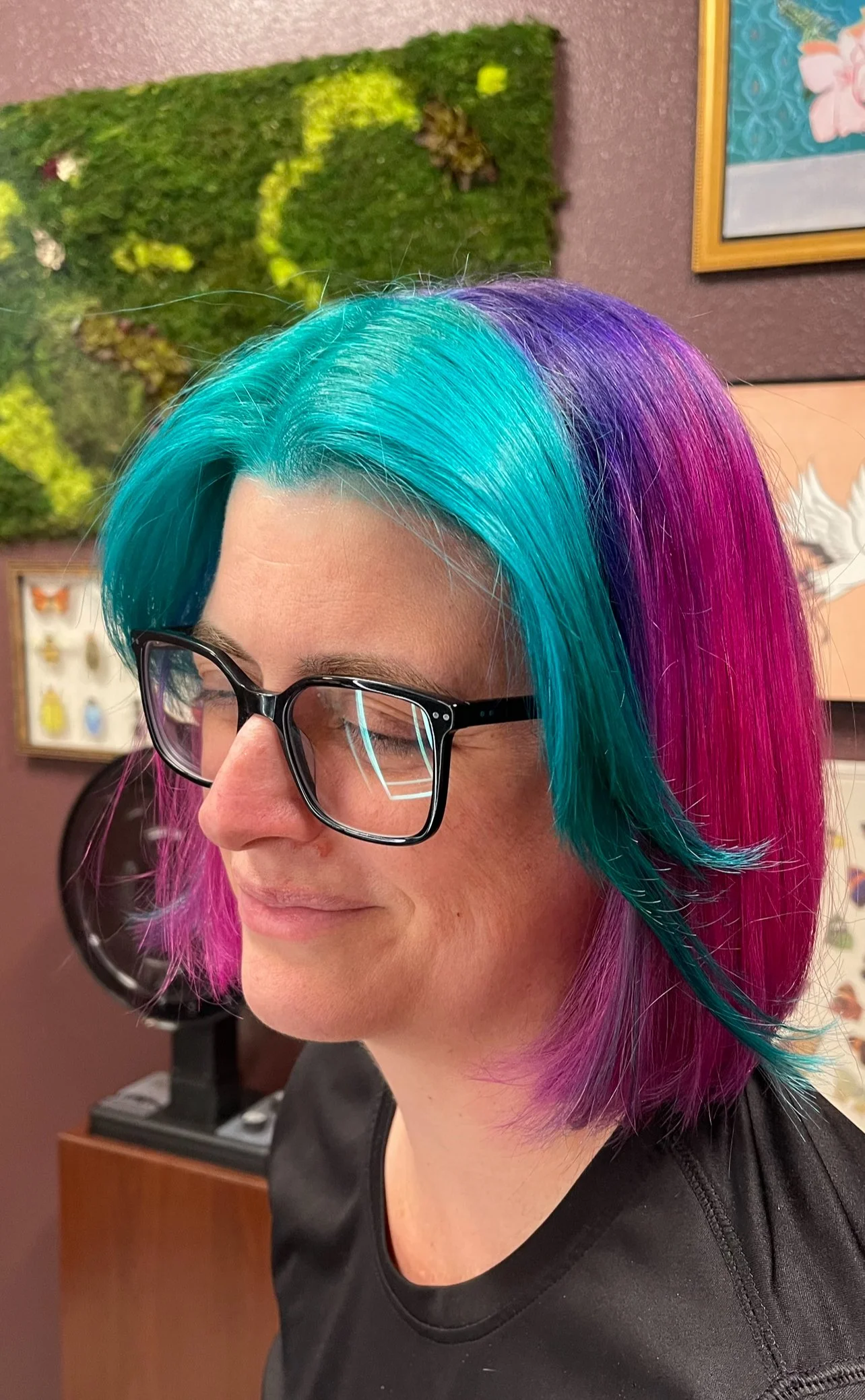 Woman with multi-colored hair dyed in shades of blue, purple, and pink, wearing black glasses, smiling with eyes closed.