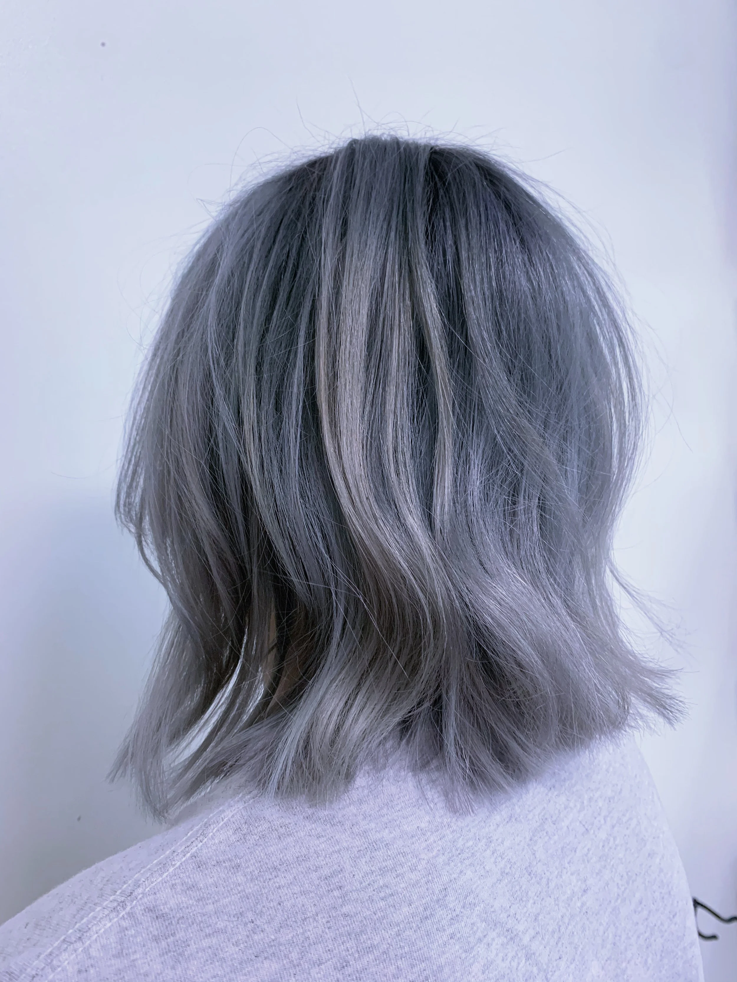 Back of a person's head with shoulder-length, wavy gray hair, wearing a light gray top, against a plain, light-colored background.