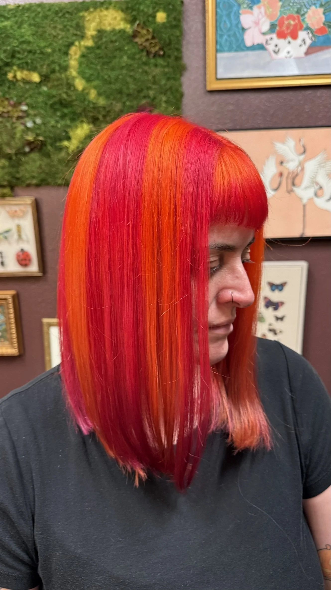 Woman with vibrant red and orange layered hair in an indoor art gallery, with framed artwork and green textured wall in the background.