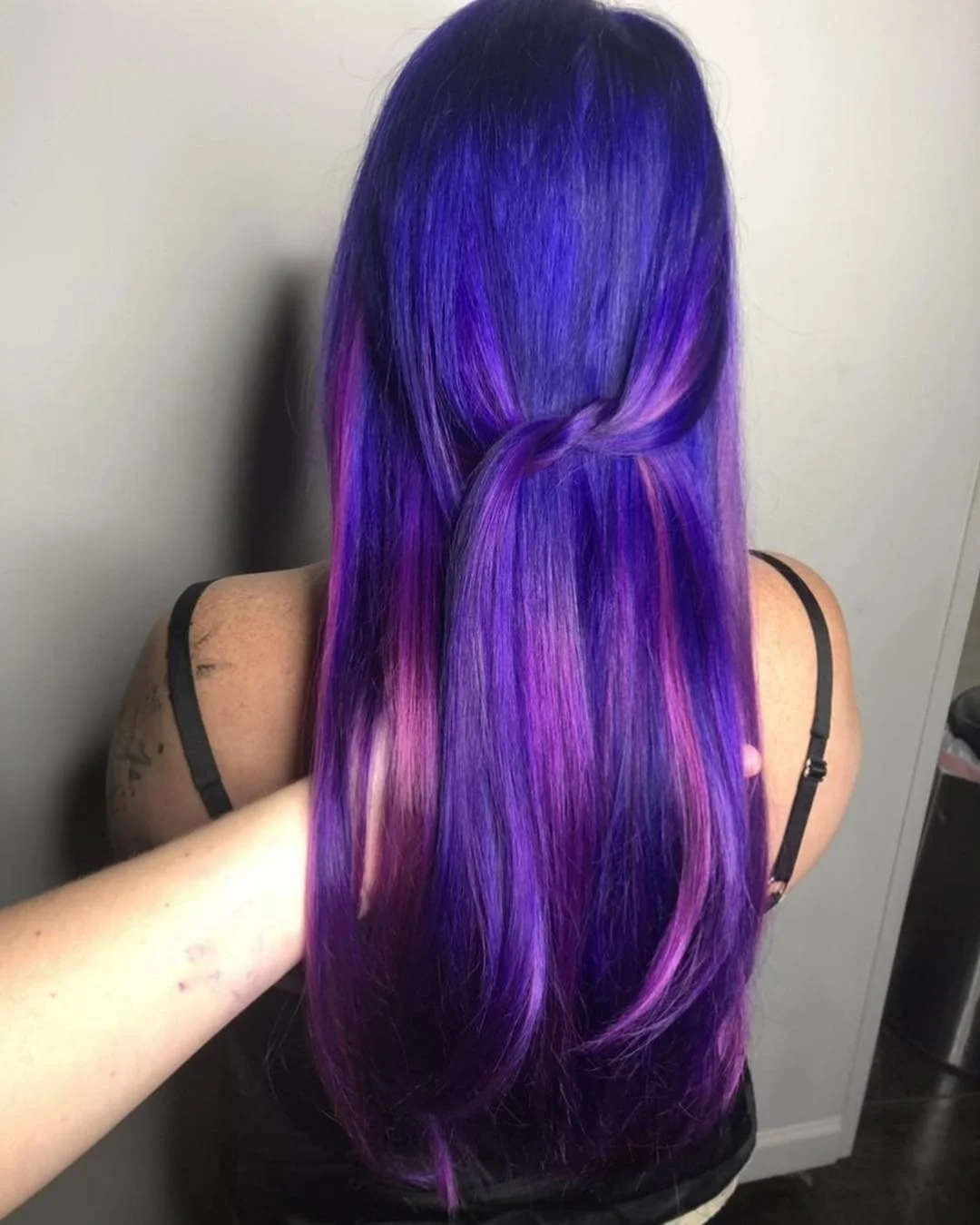 Person with long, straight, vibrant purple hair with pink highlights, styled with a small twist in the middle, wearing a black spaghetti strap top.