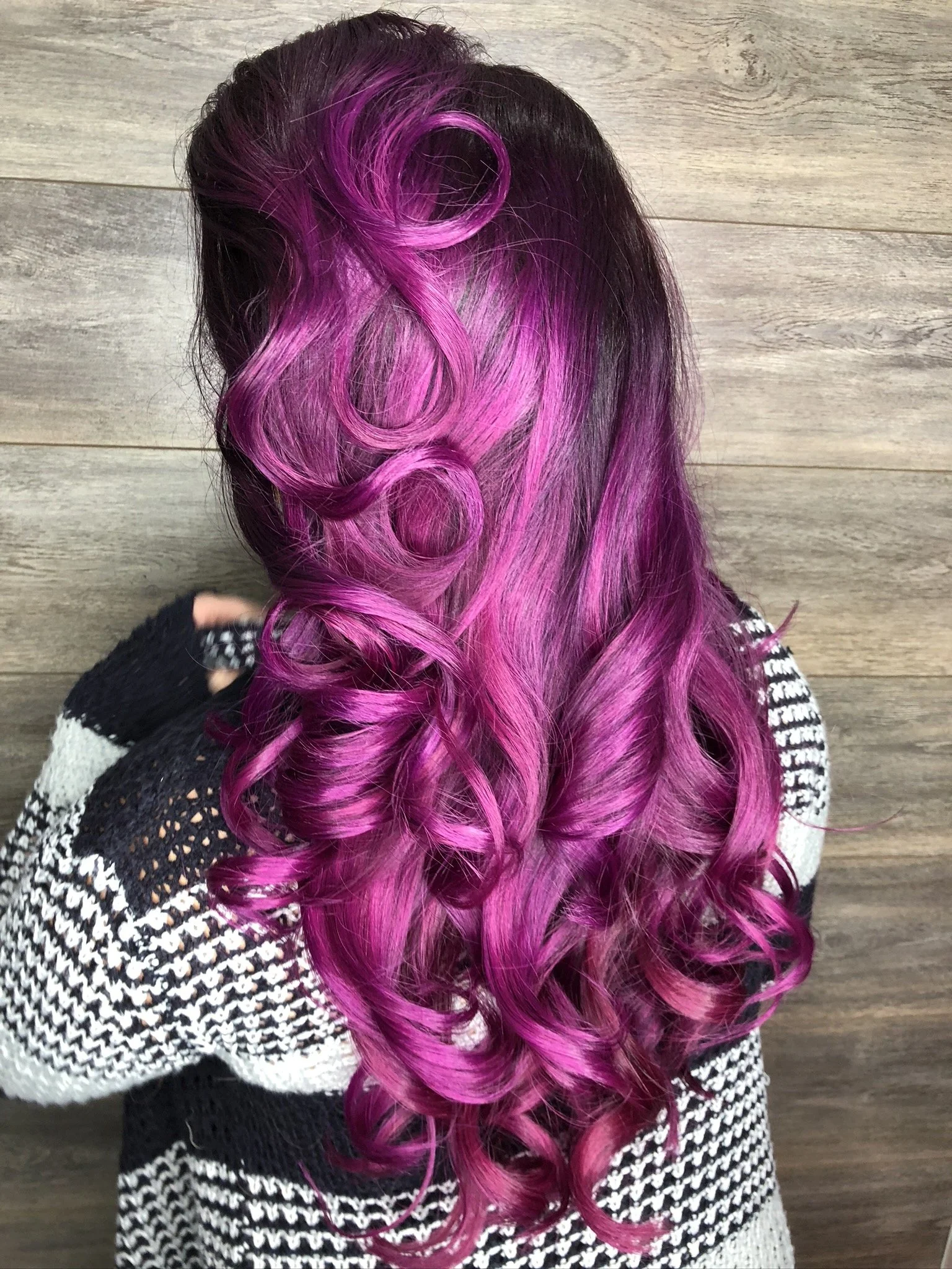 A woman with long, curly hair dyed in shades of pink and purple, with some darker roots, stands against a wooden background.