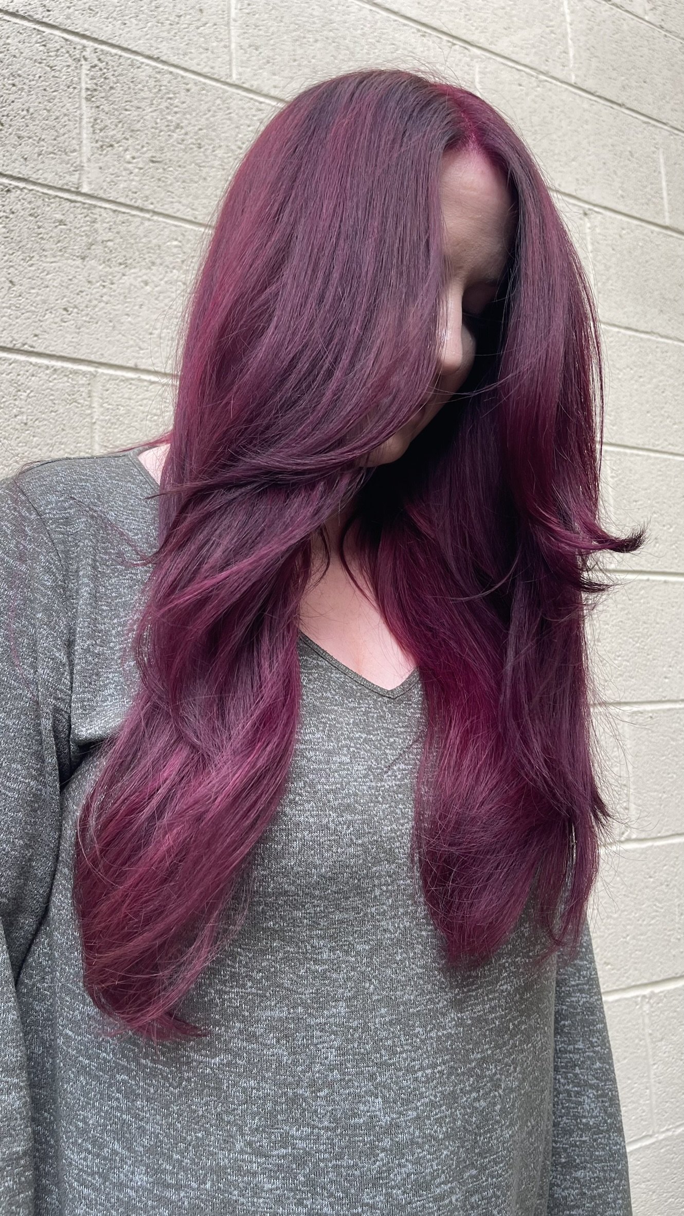 A woman with long, wavy hair dyed in shades of purple and magenta, standing against a beige brick wall, wearing a gray shirt.