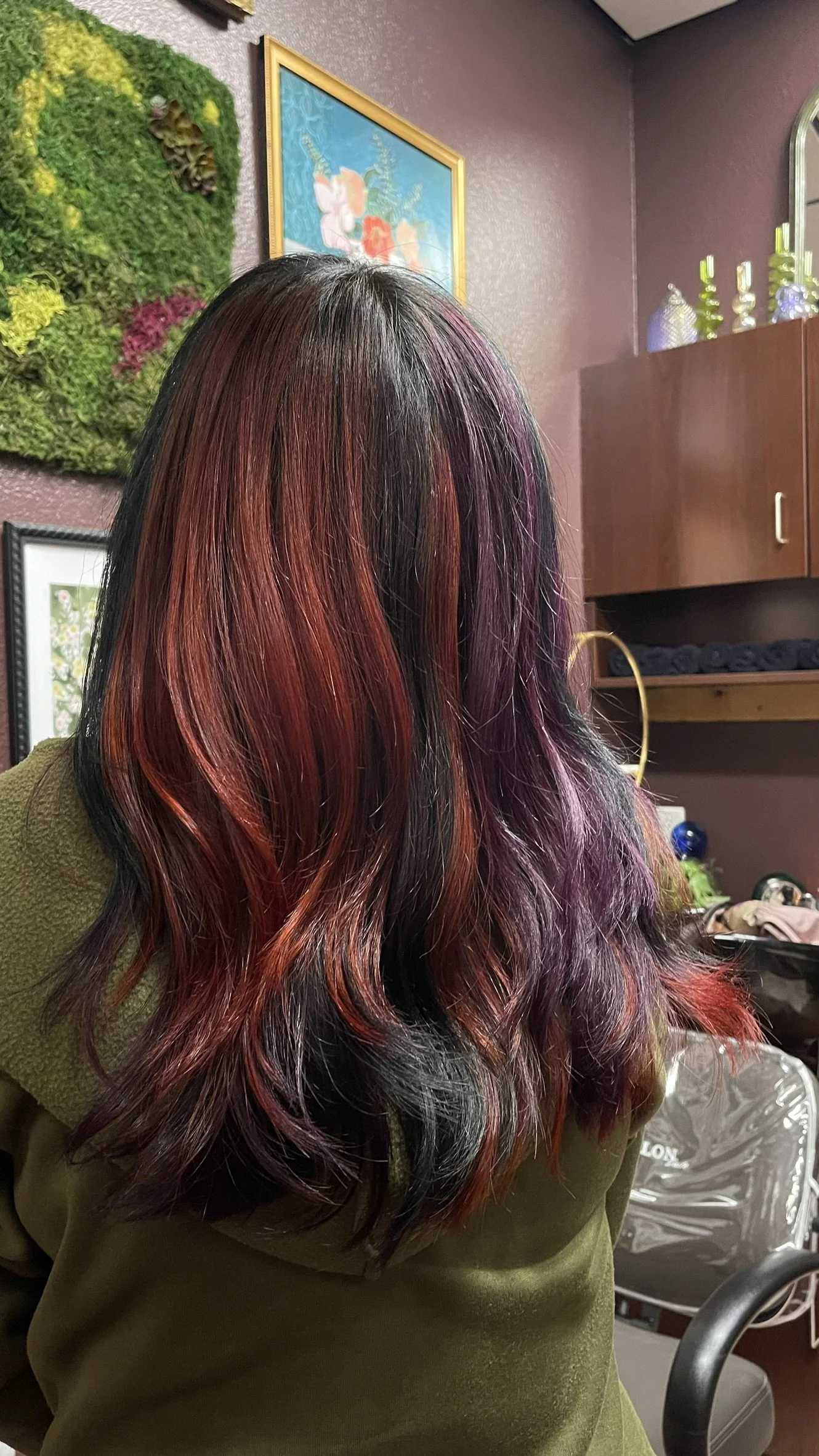 Back of a woman's head with long, wavy hair dyed in red, purple, and black shades, in a room with floral and nature-themed decorations on the wall.
