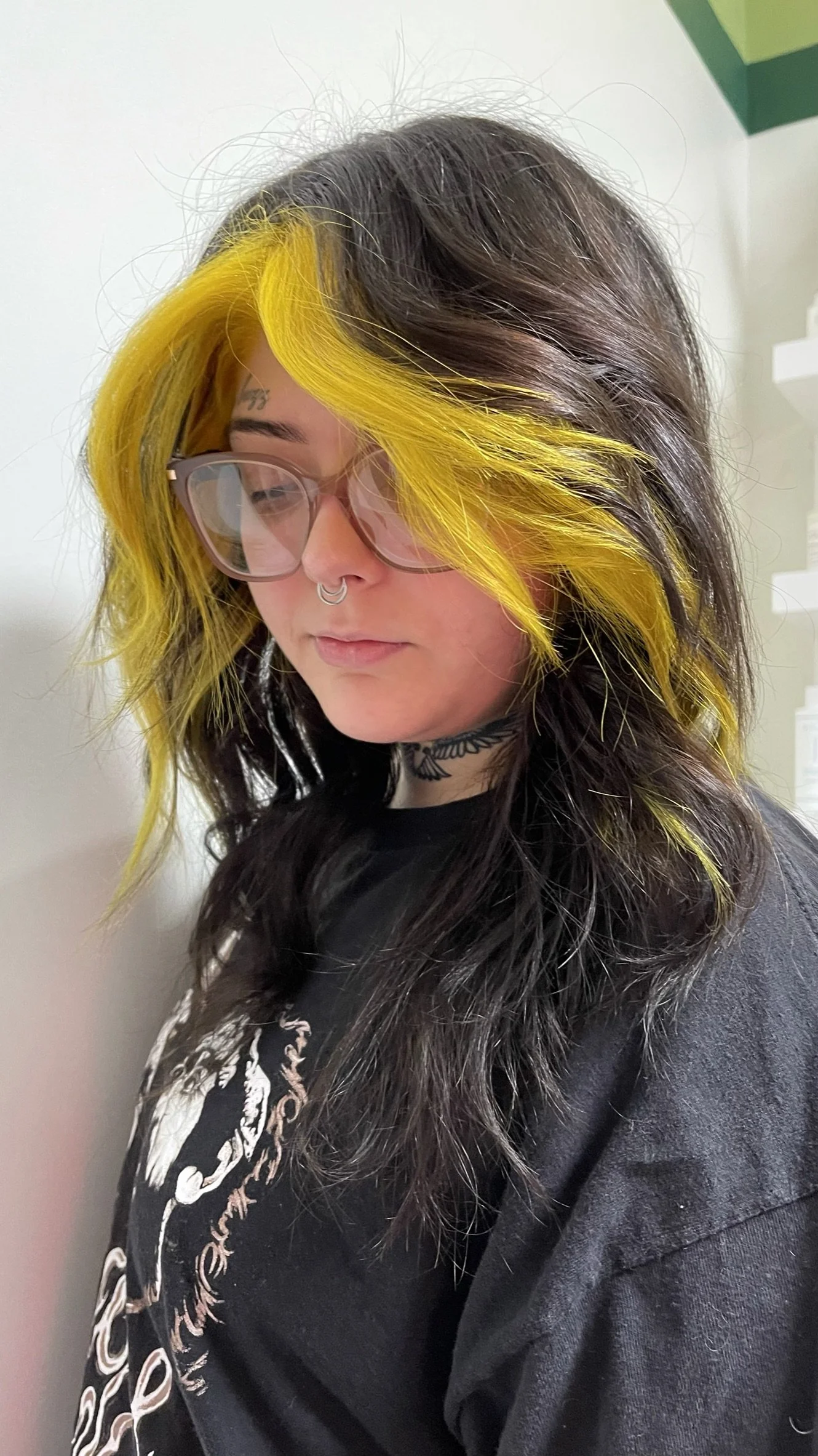 A young woman with long, dark hair and bright yellow highlights, wearing glasses, a tattoo choker, and a black T-shirt with a skeleton graphic, standing against a plain background.