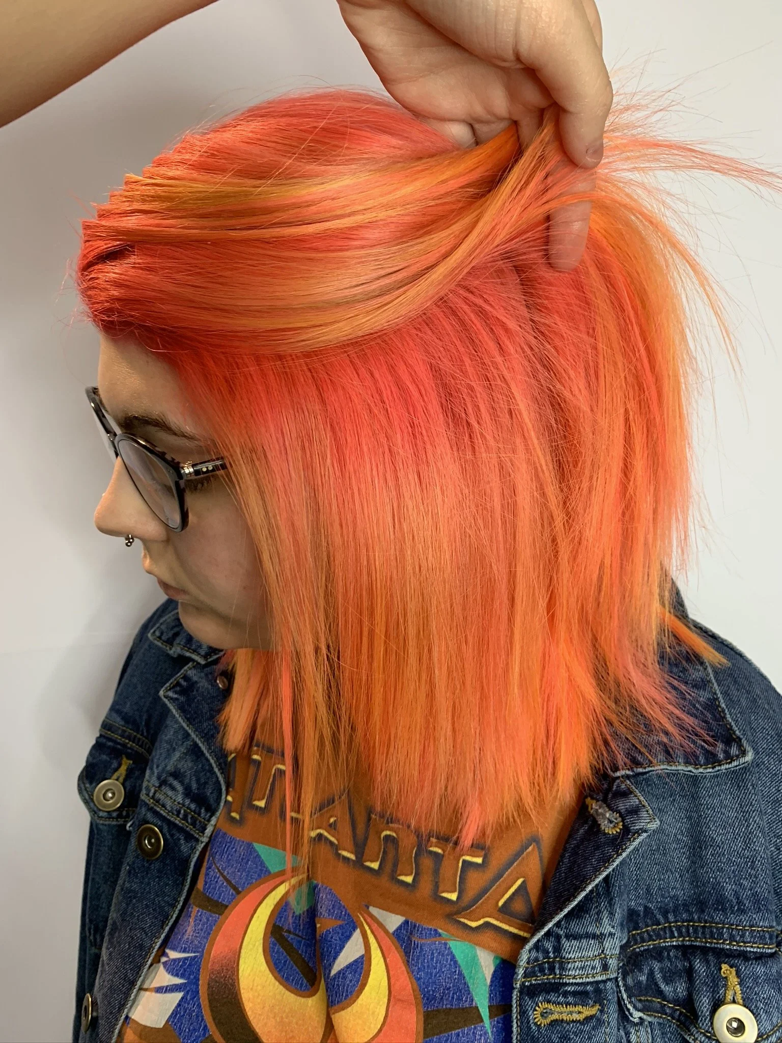 Person with bright orange hair, wearing sunglasses and a denim jacket, viewed from the side.