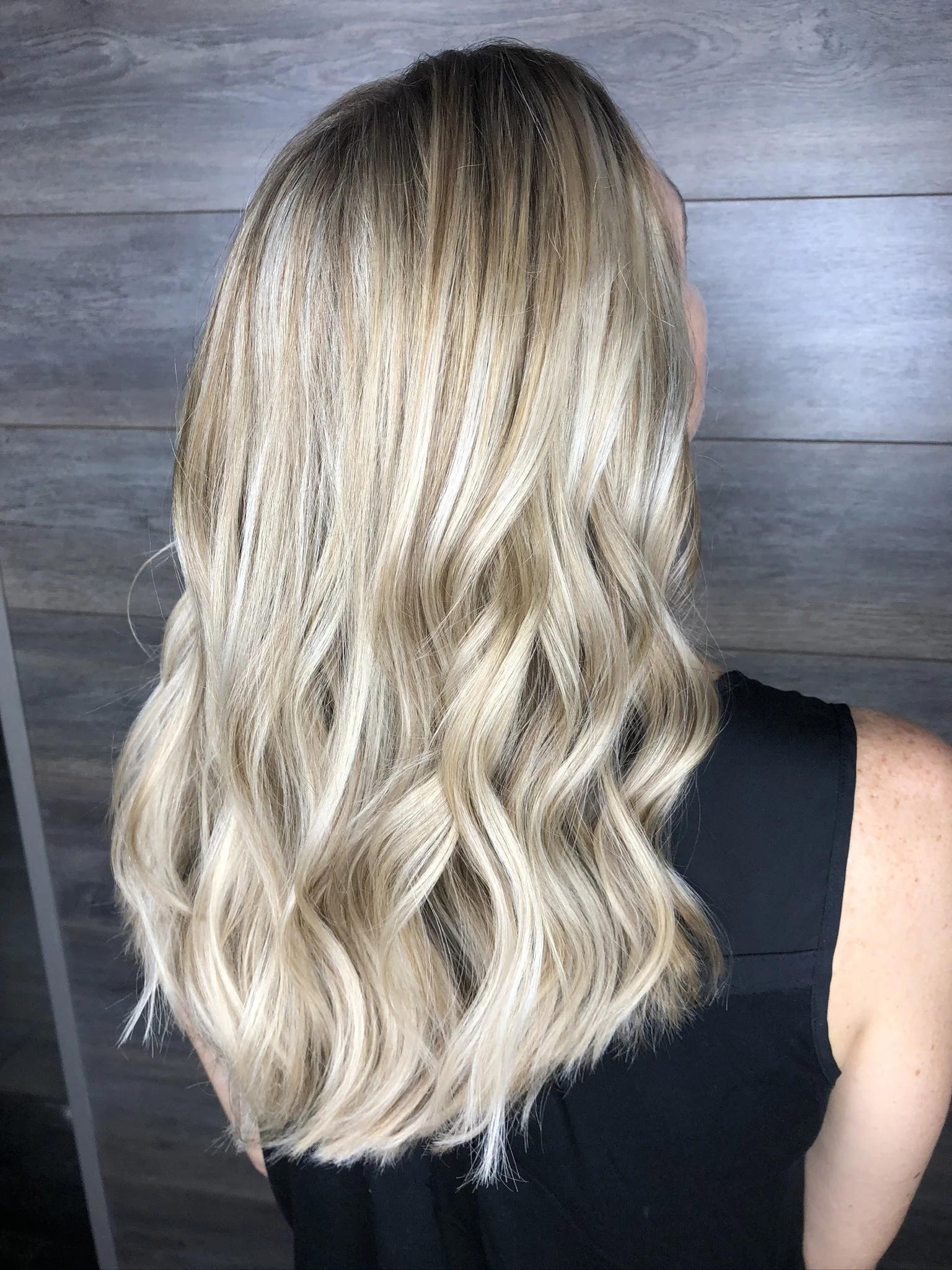 Back view of a woman with long, curly blonde hair wearing a black sleeveless top, standing against a wooden wall.