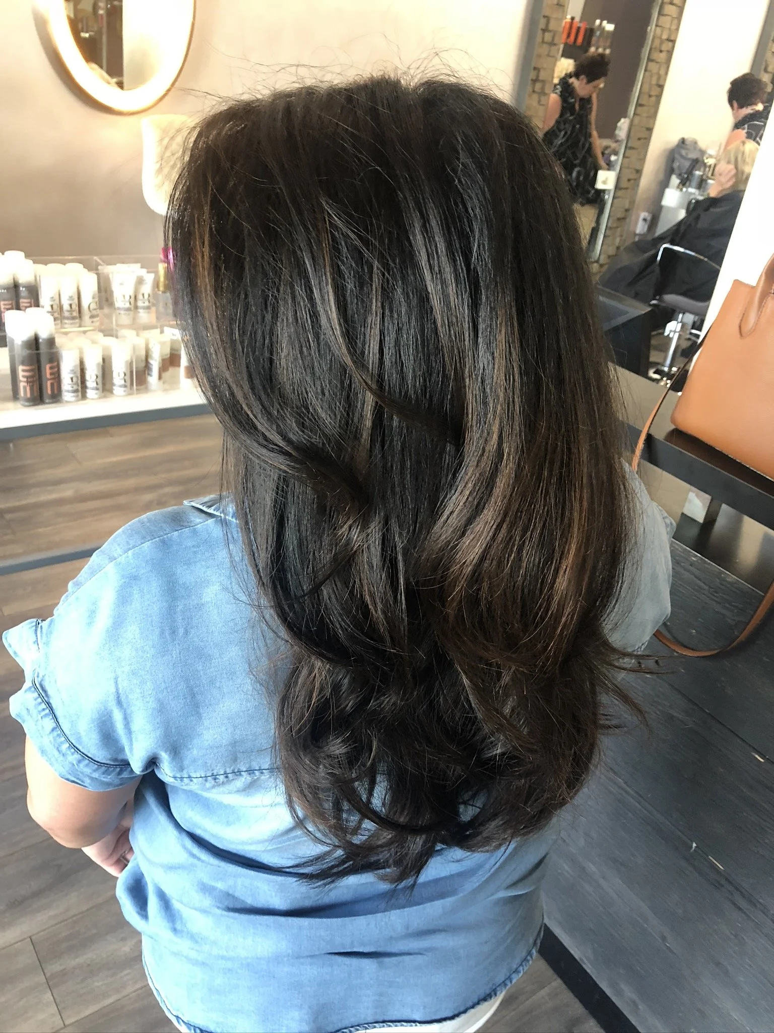 A woman with dark brown hair styled in loose waves, wearing a light blue denim shirt, sitting in a salon.