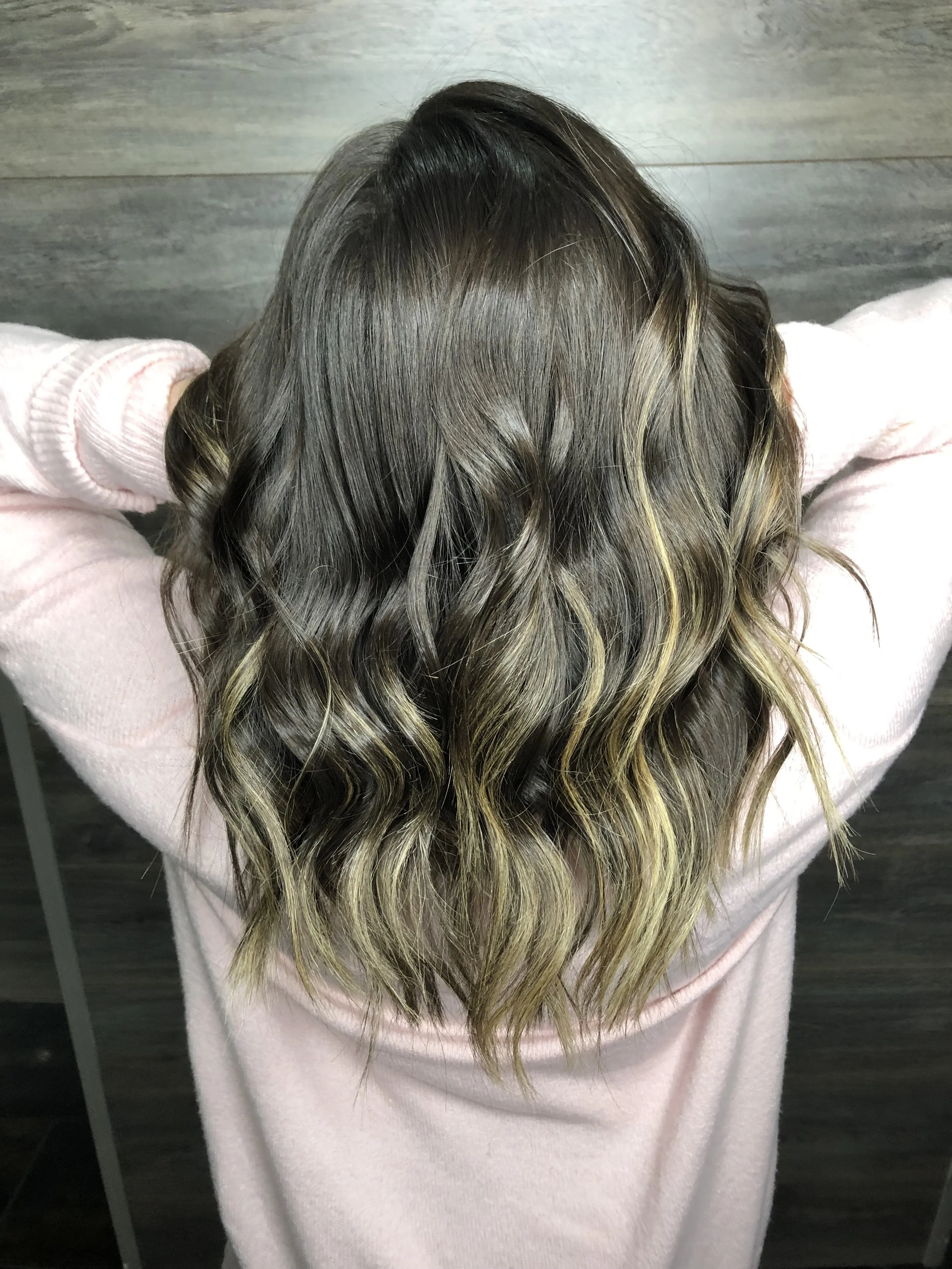 A woman with long, wavy brown hair with blonde highlights, viewed from above, wearing a light pink sweater.