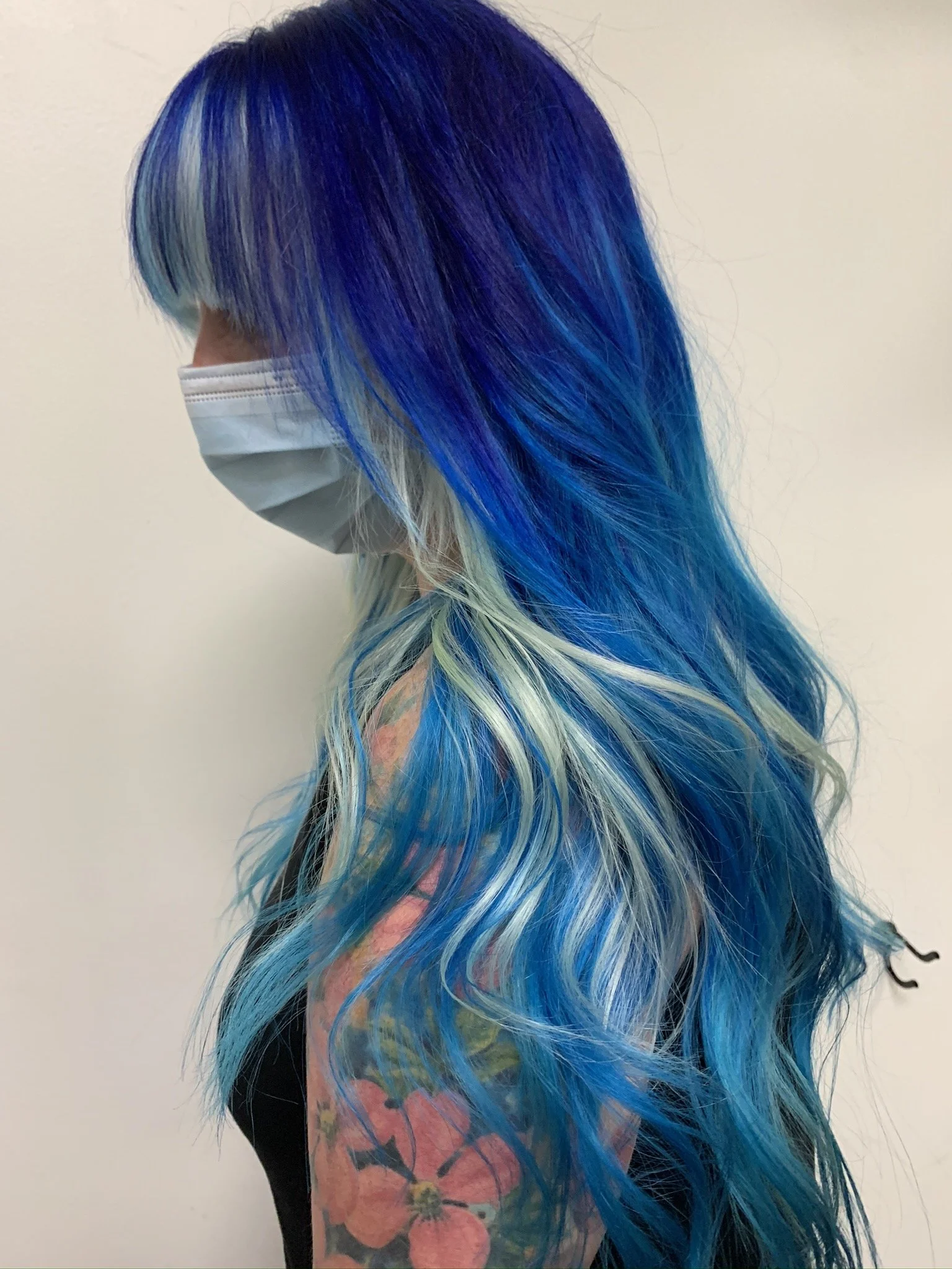 Side view of a woman with long, wavy hair styled in vibrant shades of blue, turquoise, and purple, wearing a face mask.