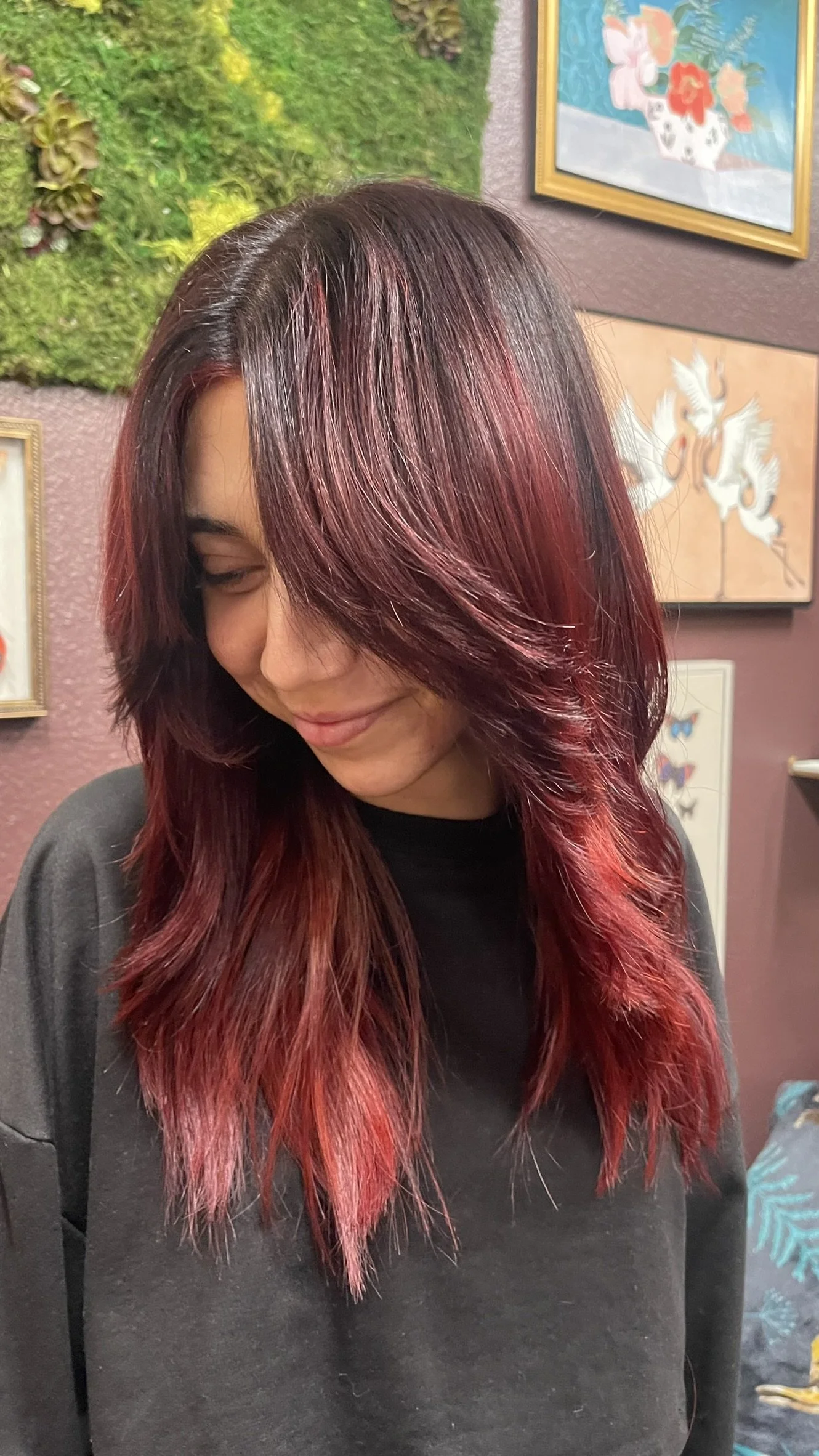 A woman with shoulder-length dark hair with red highlights, smiling and looking down, standing indoors with colorful artwork on the walls behind her.