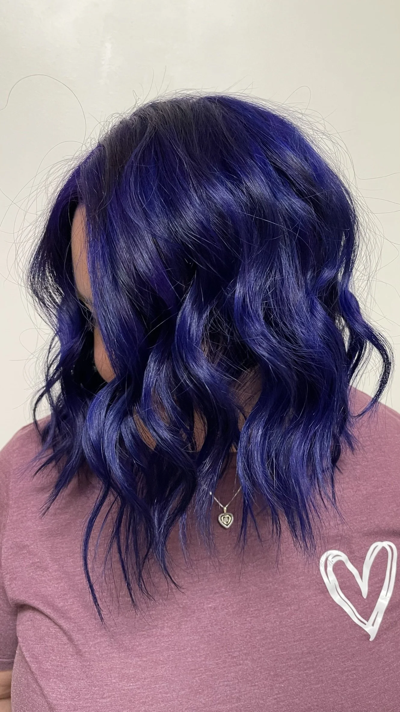 Woman with shoulder-length wavy hair dyed in shades of purple and blue, wearing a pink shirt with a white heart drawing and a silver necklace with a heart pendant.
