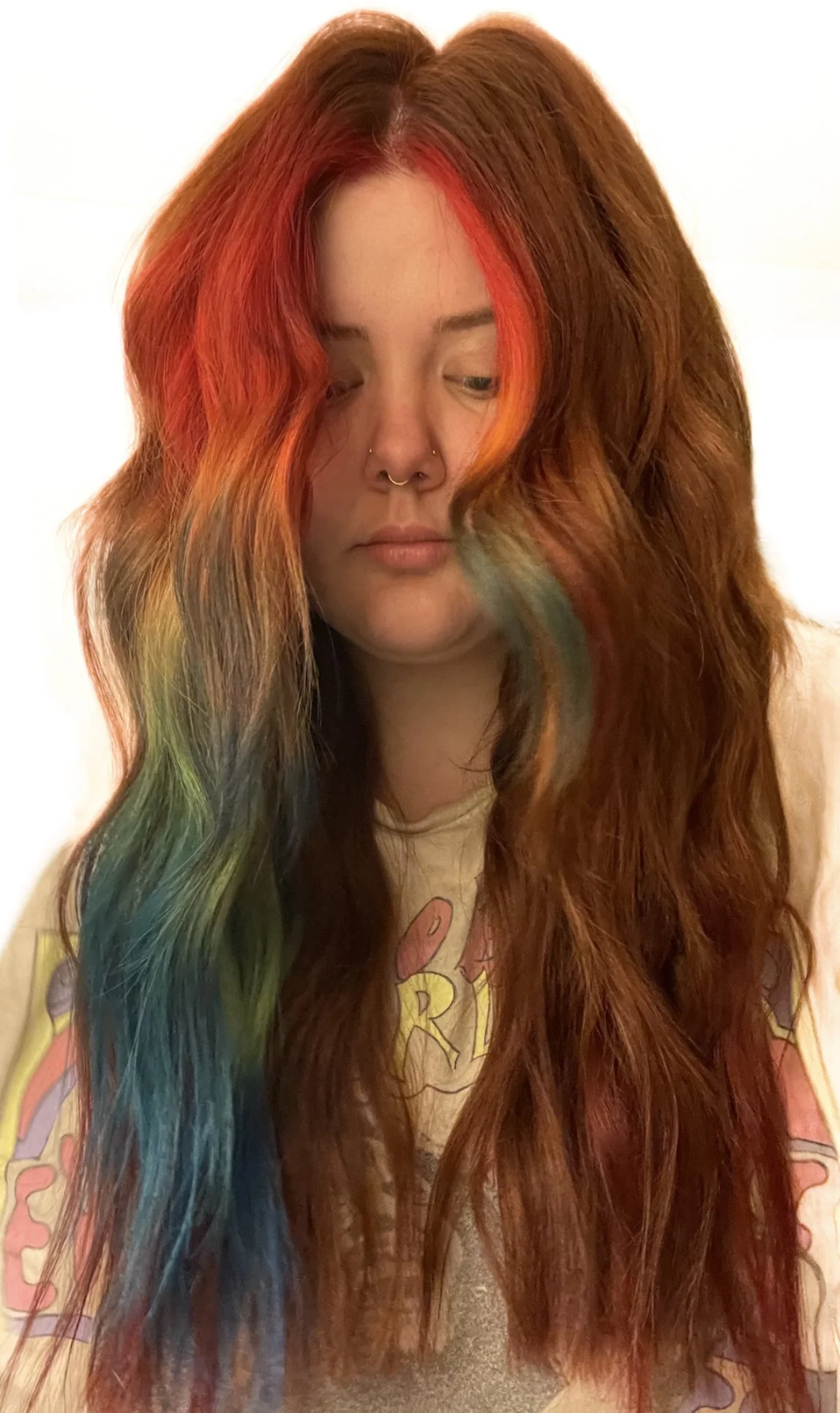 A woman with long, wavy red hair featuring rainbow streaks, looking down with a neutral expression. She has light skin, multiple facial piercings, and is wearing a multicolored hoodie with yellow writing.