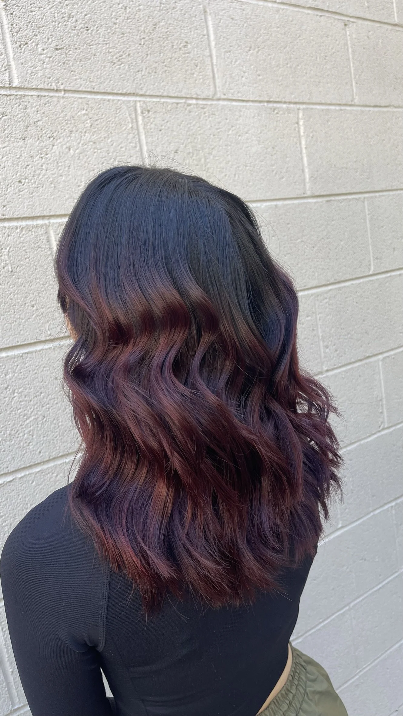A woman with wavy hair dyed in an ombre style with black roots that fade into purple and red tips, standing against a white brick wall.