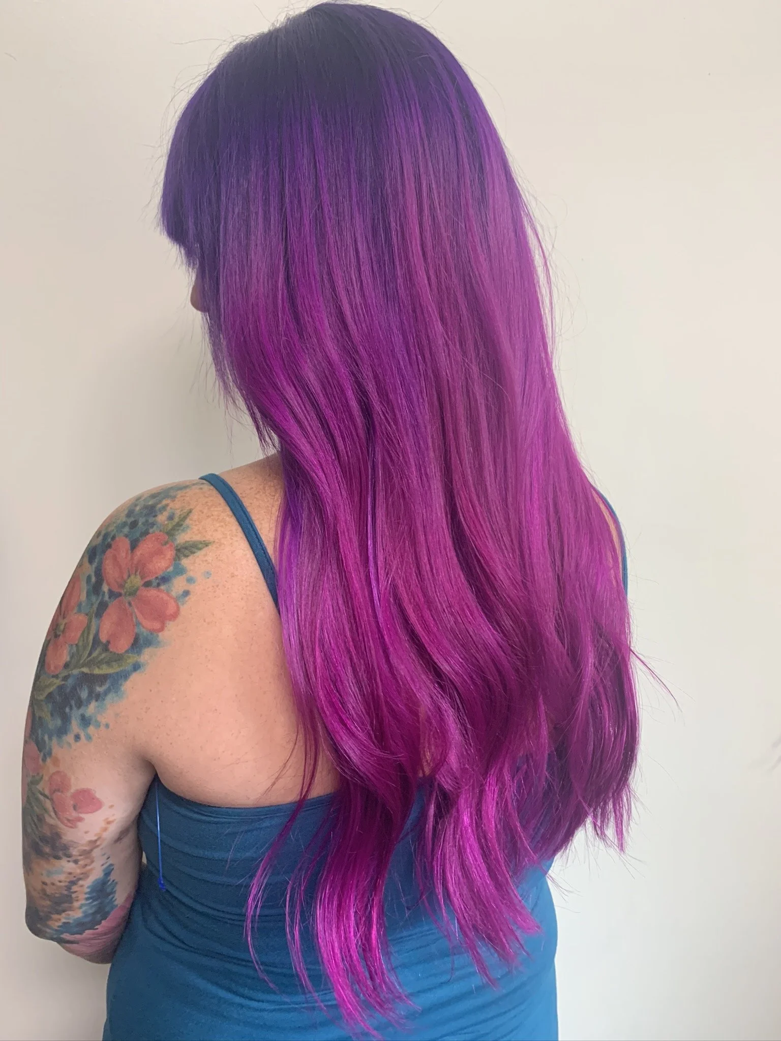 Woman with long, vibrant purple and pink hair, wearing a blue tank top, standing against a plain white wall.