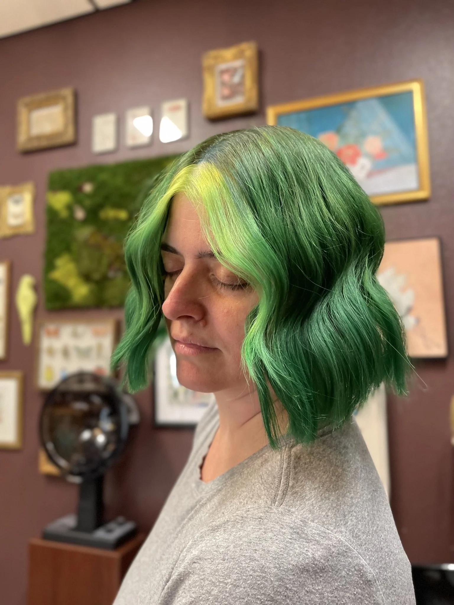 Woman with shoulder-length green hair with a yellow streak, wearing a gray shirt, standing against a wall decorated with framed artwork.