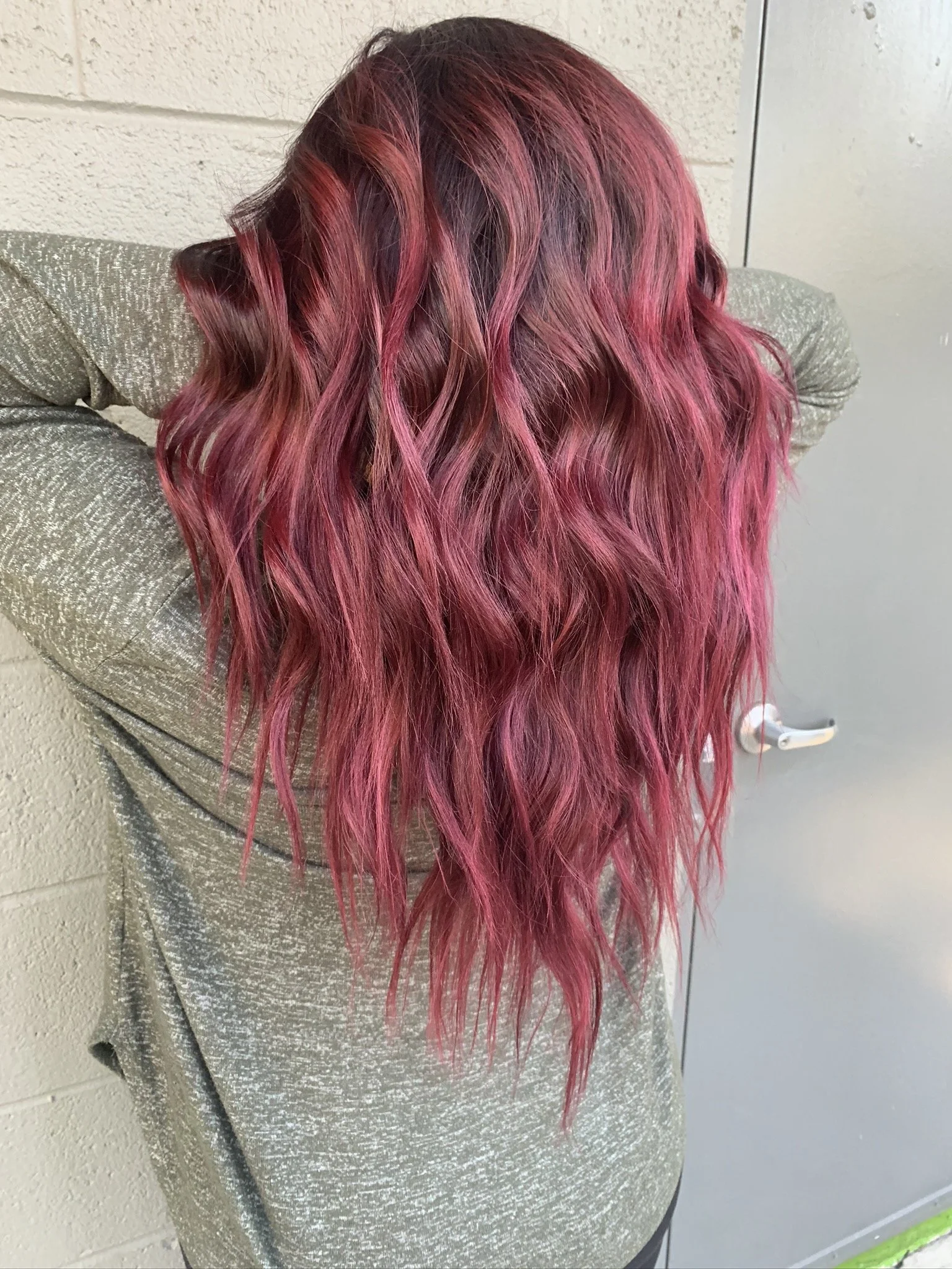 Back view of a person with long, wavy hair dyed in shades of pink and red, wearing a light-colored, textured sweater.
