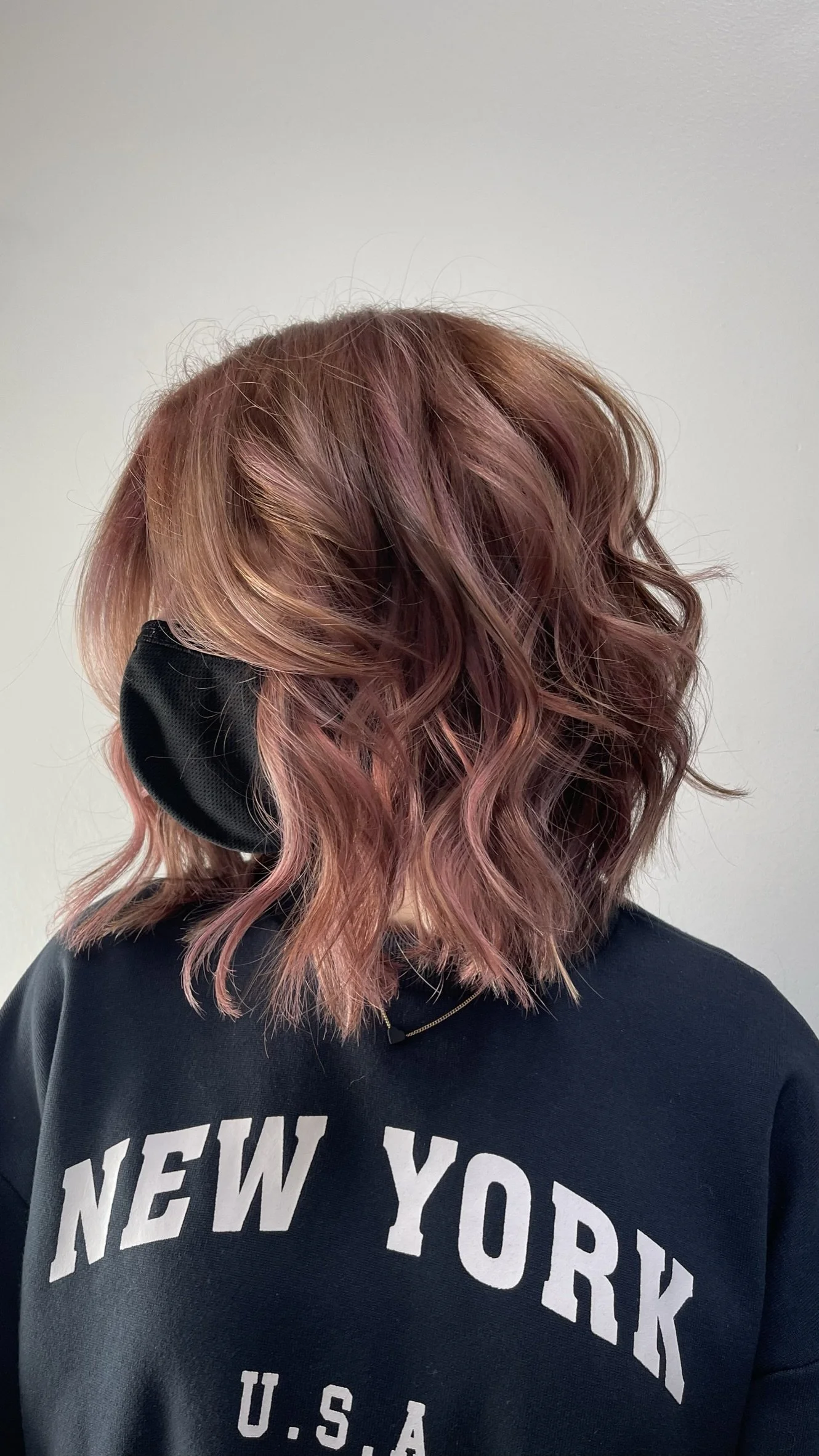 Side view of a person with shoulder-length wavy hair that is dyed in shades of pink and auburn, wearing a black face mask and a navy sweatshirt with "NEW YORK" printed on it.