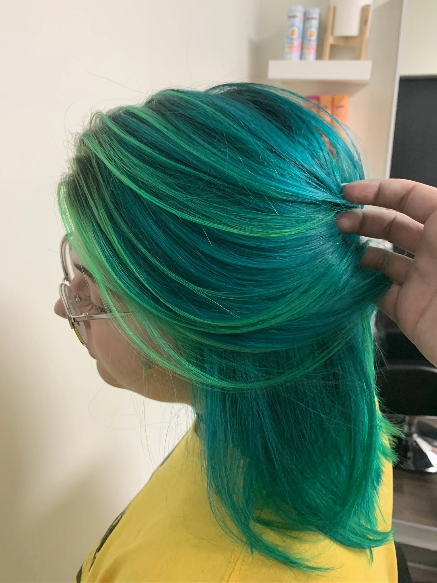 Woman with vibrant turquoise and green layered hair, wearing glasses and a yellow top, holding her hair with one hand.