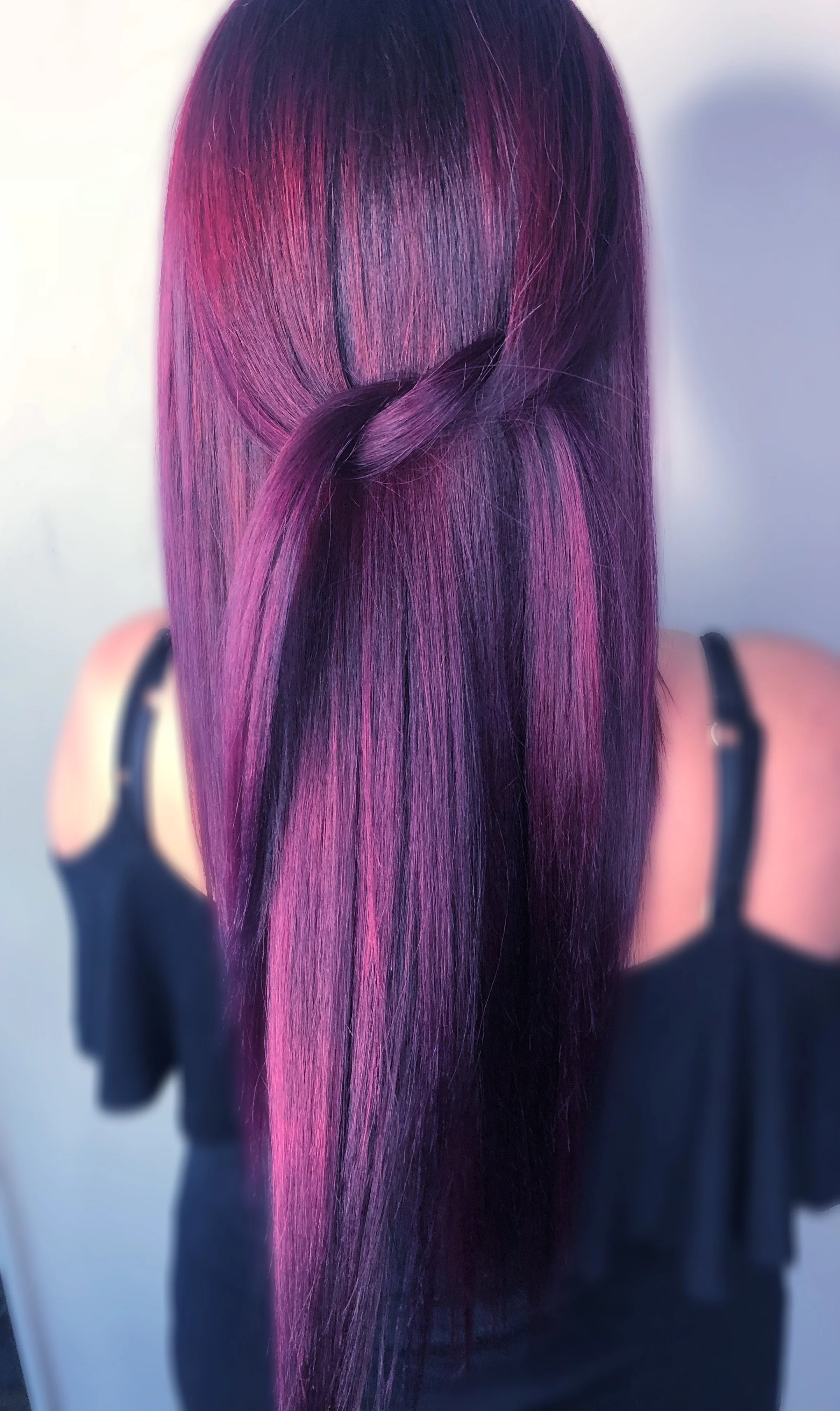 Back view of a woman with long, straight, purple and pink ombre hair styled in a loop knot at the crown, wearing a black top with spaghetti straps.
