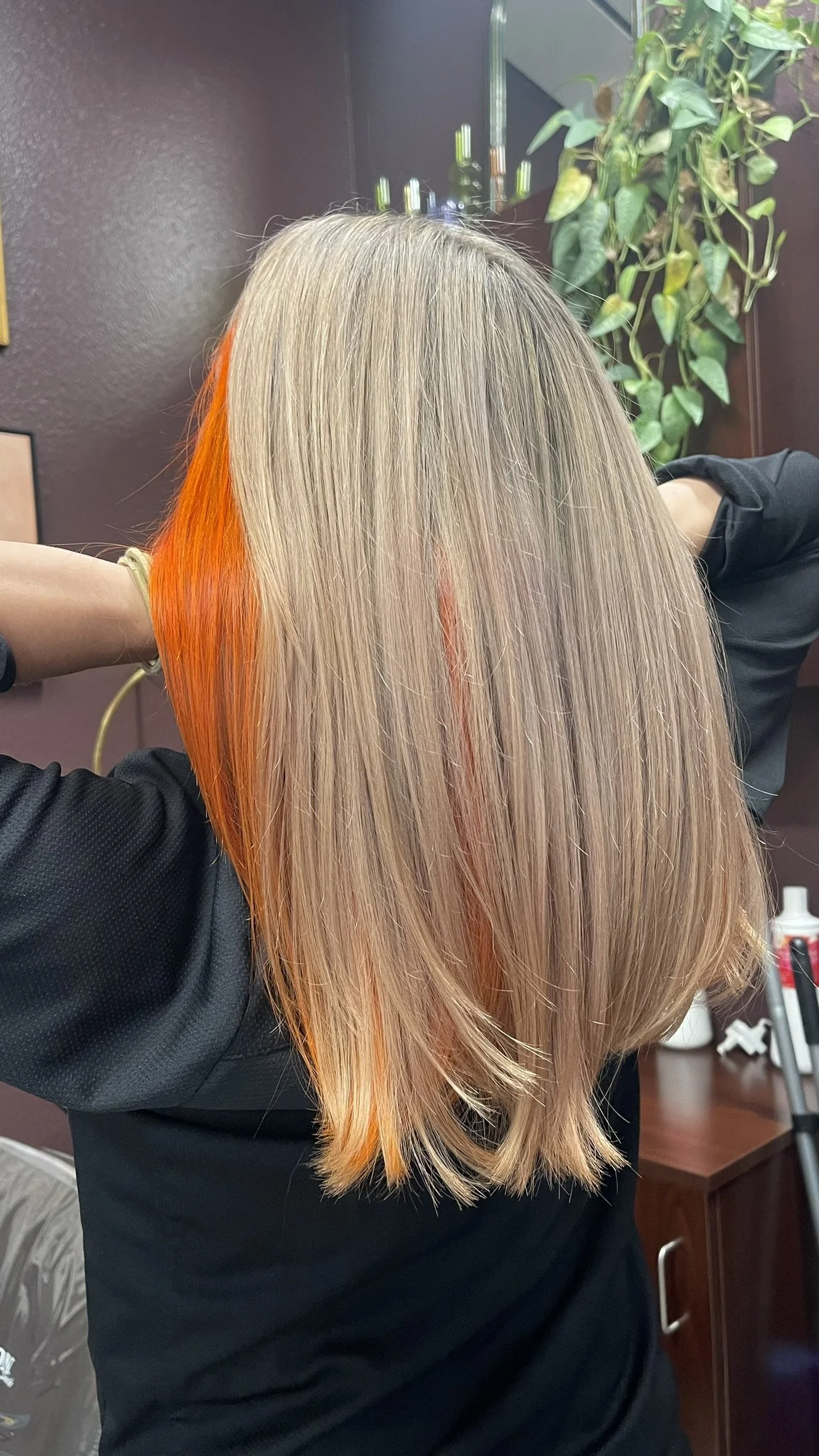 Person with shoulder-length blonde hair getting a vibrant orange streak added, seen from behind in a hair salon.