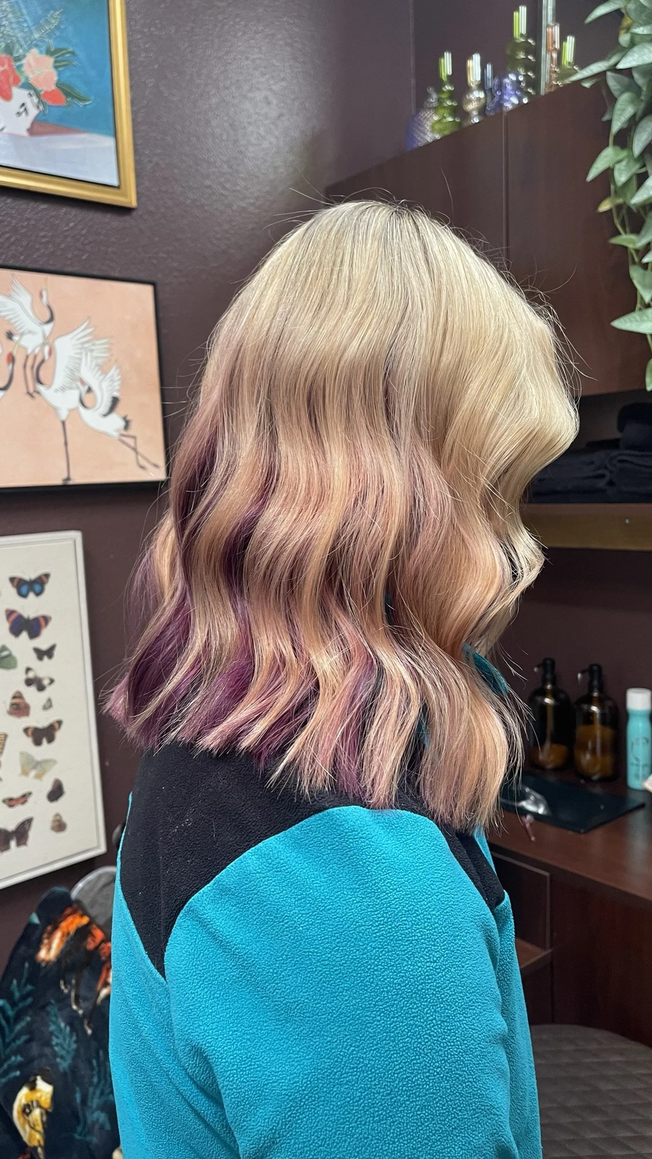 Side profile of woman with shoulder-length wavy hair styled with a blonde base and pink and purple ombre ends, wearing a blue and black jacket inside a room with butterfly artwork and decorative items.