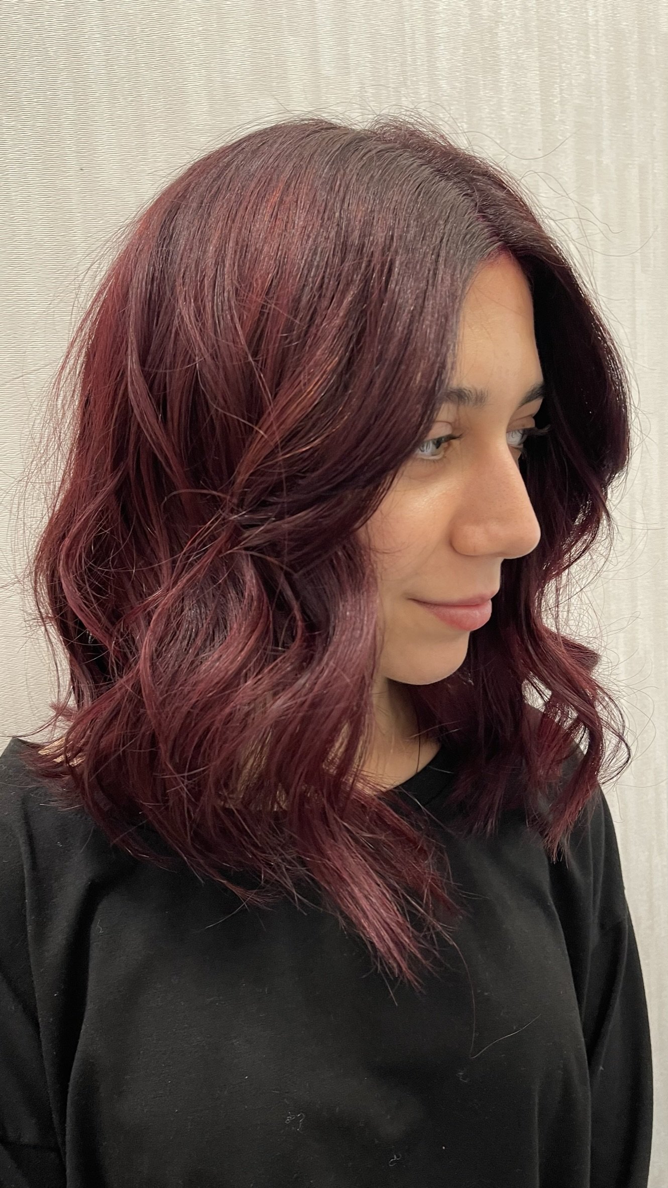 A woman with shoulder-length wavy burgundy hair, wearing a black top, standing in front of a light-colored textured wall.