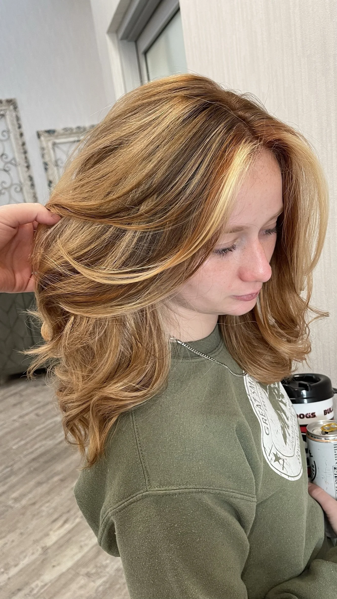 A woman with light red hair styled in loose waves, having her hair gently lifted by someone else in an indoor setting.