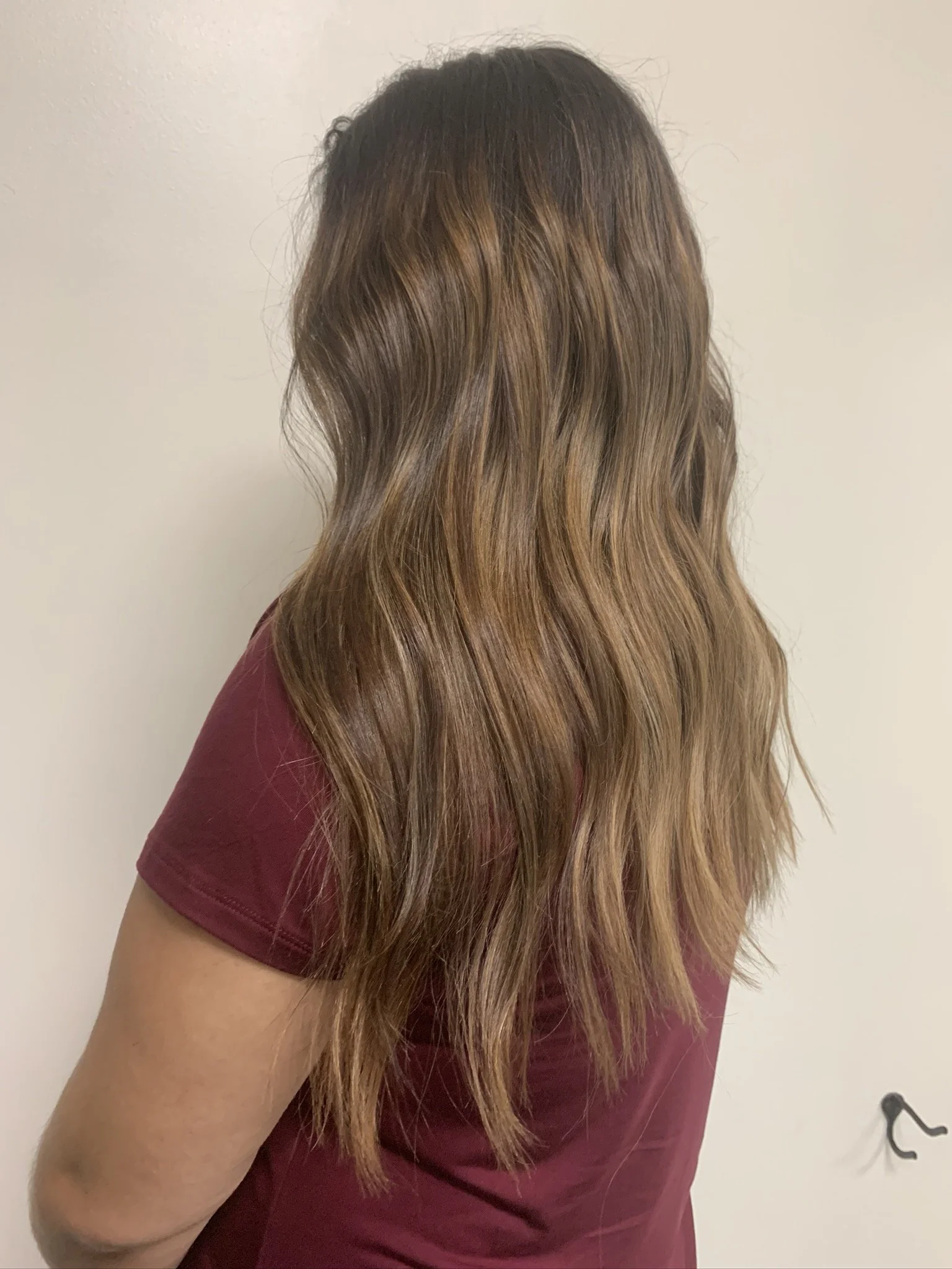 Back view of a woman with long, wavy brown hair wearing a maroon shirt against a plain wall.