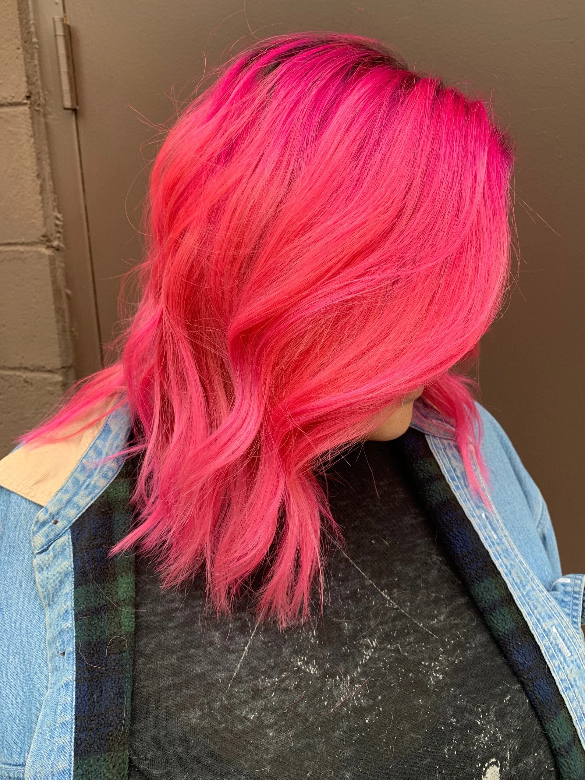 Person with bright pink, wavy hair wearing a black shirt and denim jacket, standing against a gray wall.
