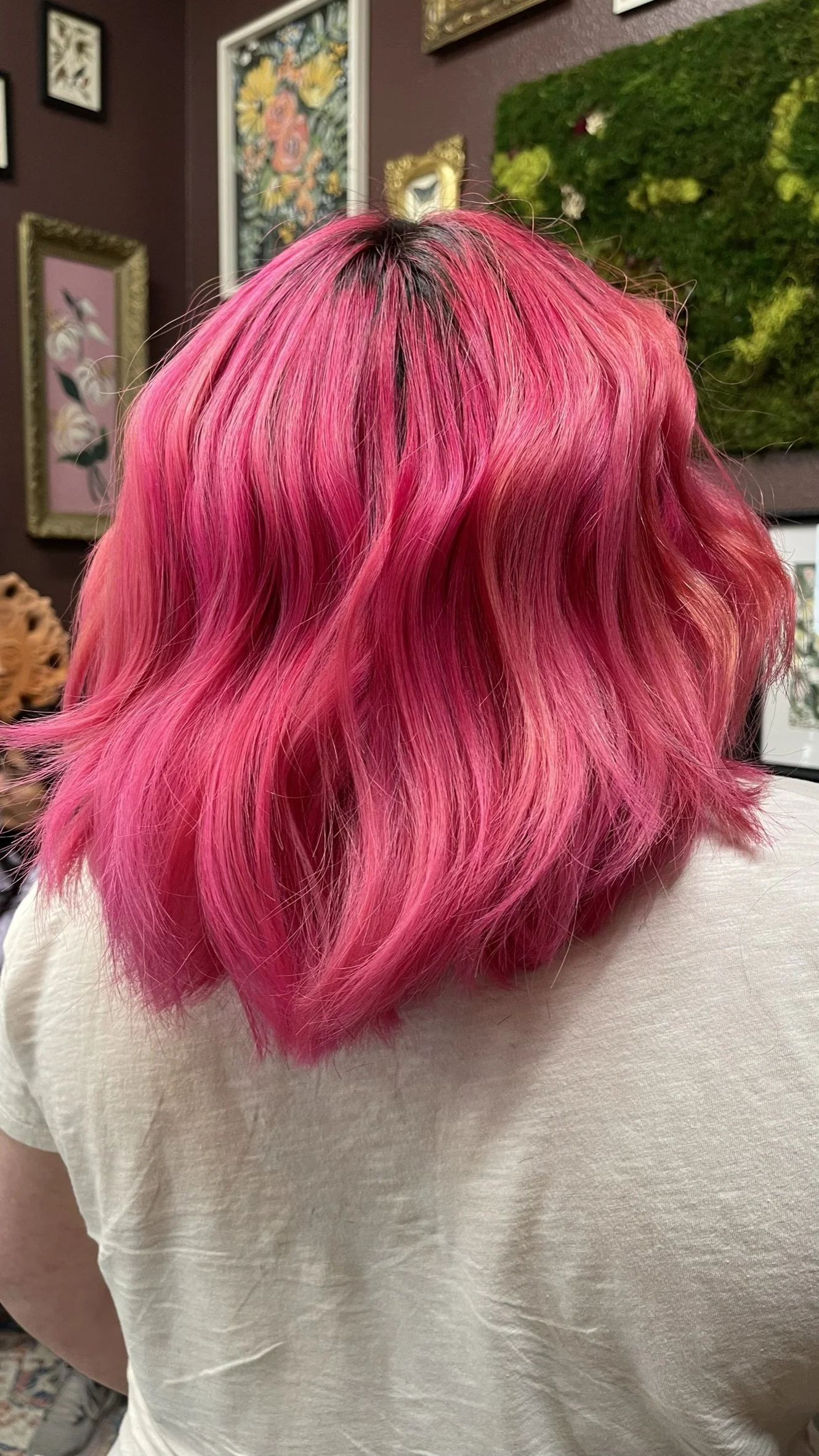 Back view of a person with short, wavy, bright pink hair. The background features framed artwork and a textured green wall.
