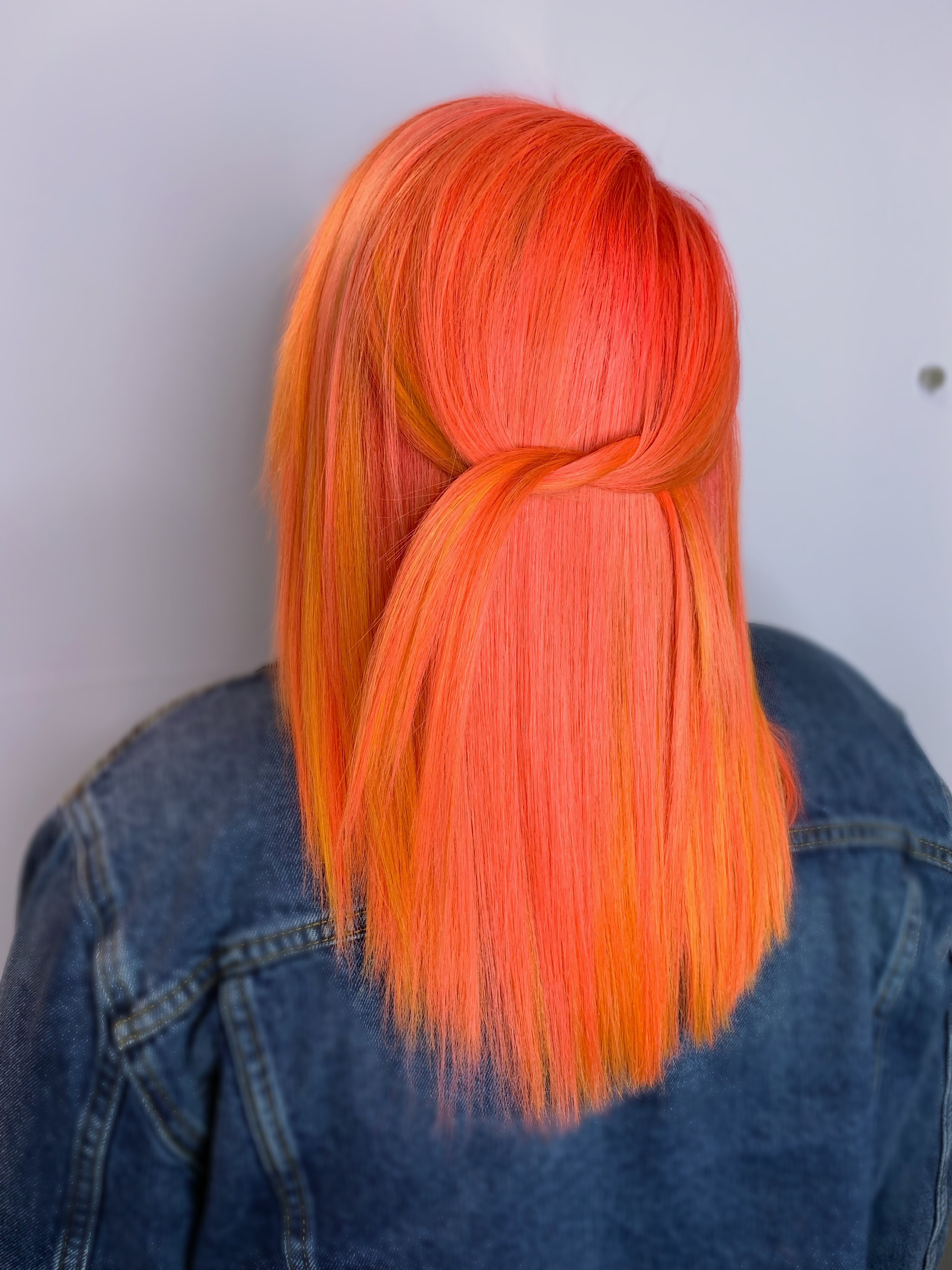Back of a person with long, straight hair dyed in vibrant shades of orange, pink, and yellow, wearing a denim jacket.